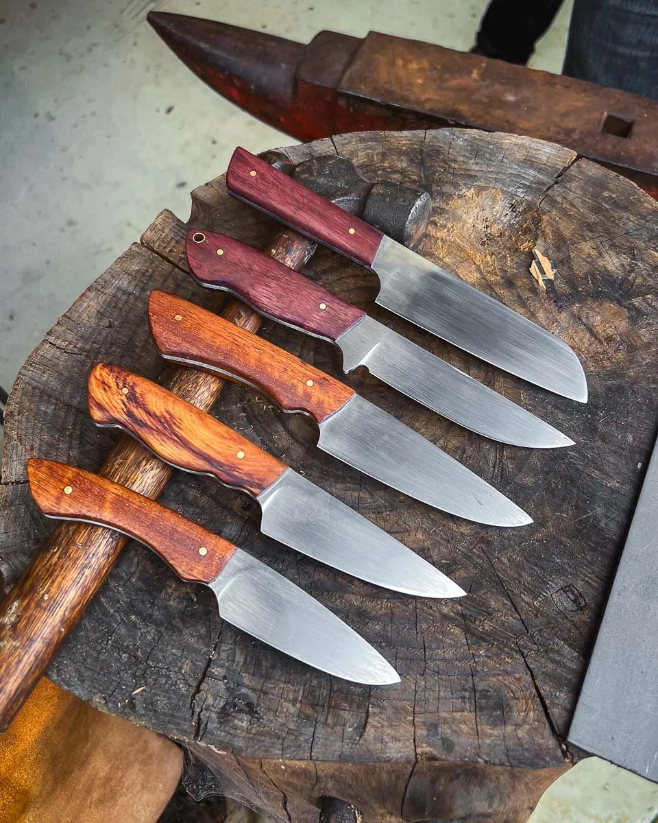 Four handcrafted kitchen knives with different colored wooden handles, placed on a wooden chopping block, with a hammer and an anvil nearby.