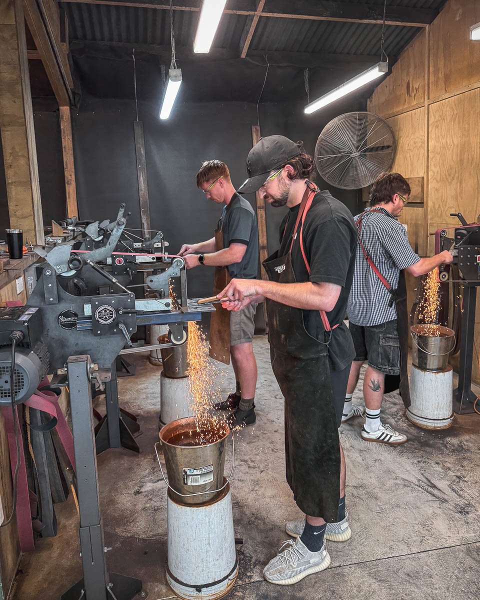 Three men are working in a workshop, each using a metalworking machine that produces sparks. The environment has a rustic, industrial feel with wooden walls and exposed ceiling beams.