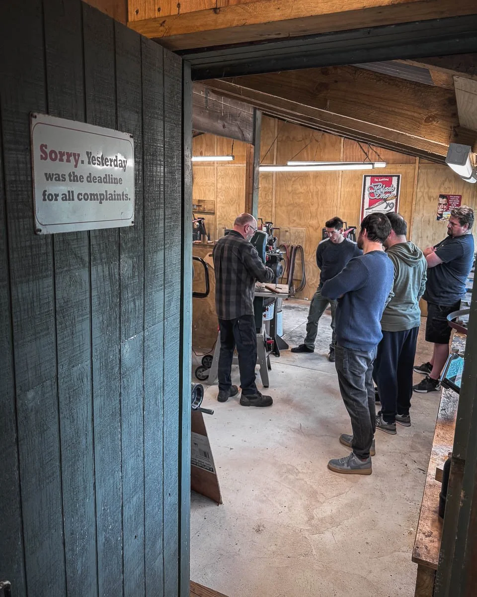 A group of five men standing around a woodwork machine in a workshop, listening to an instructor demonstrate.
