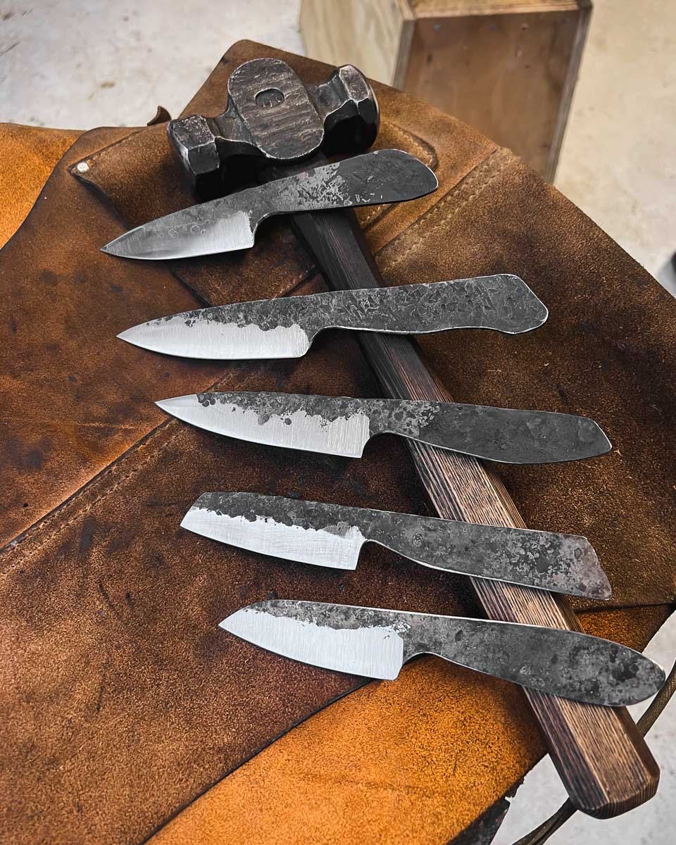 Five hand-forged knives with rustic, textured blades and a small black sharpening tool on a worn leather apron.