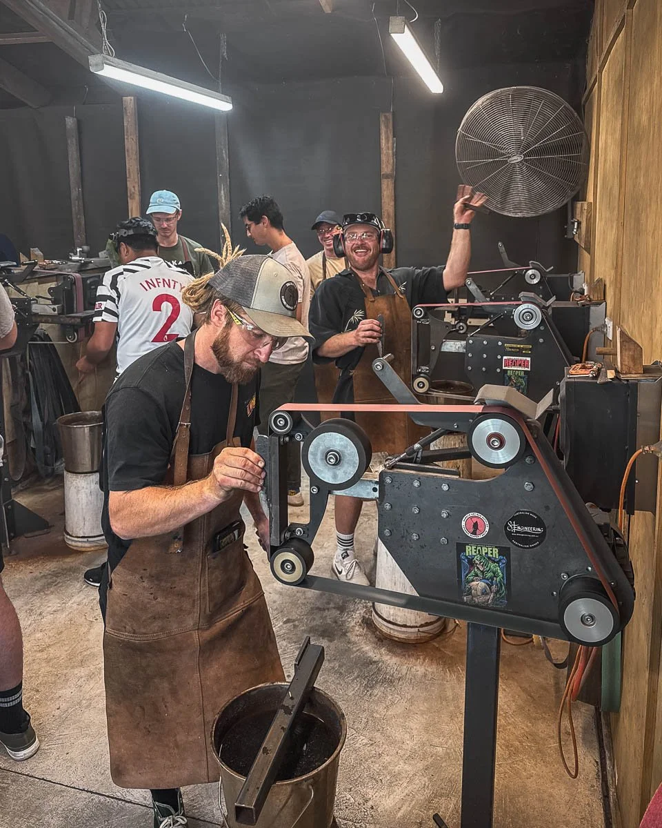 People working in a woodworking shop, one man is operating a machine with a belt and pulley system, another man is checking something on the machine, with others in the background observing and chatting.