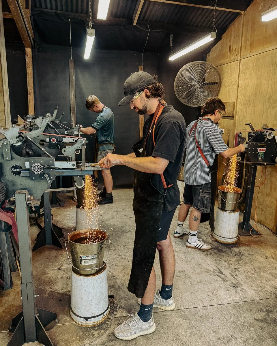 Three men working in a workshop, grinding metal with sparks flying into buckets. The room has a black wall, wooden walls, and a ceiling with fluorescent lights.