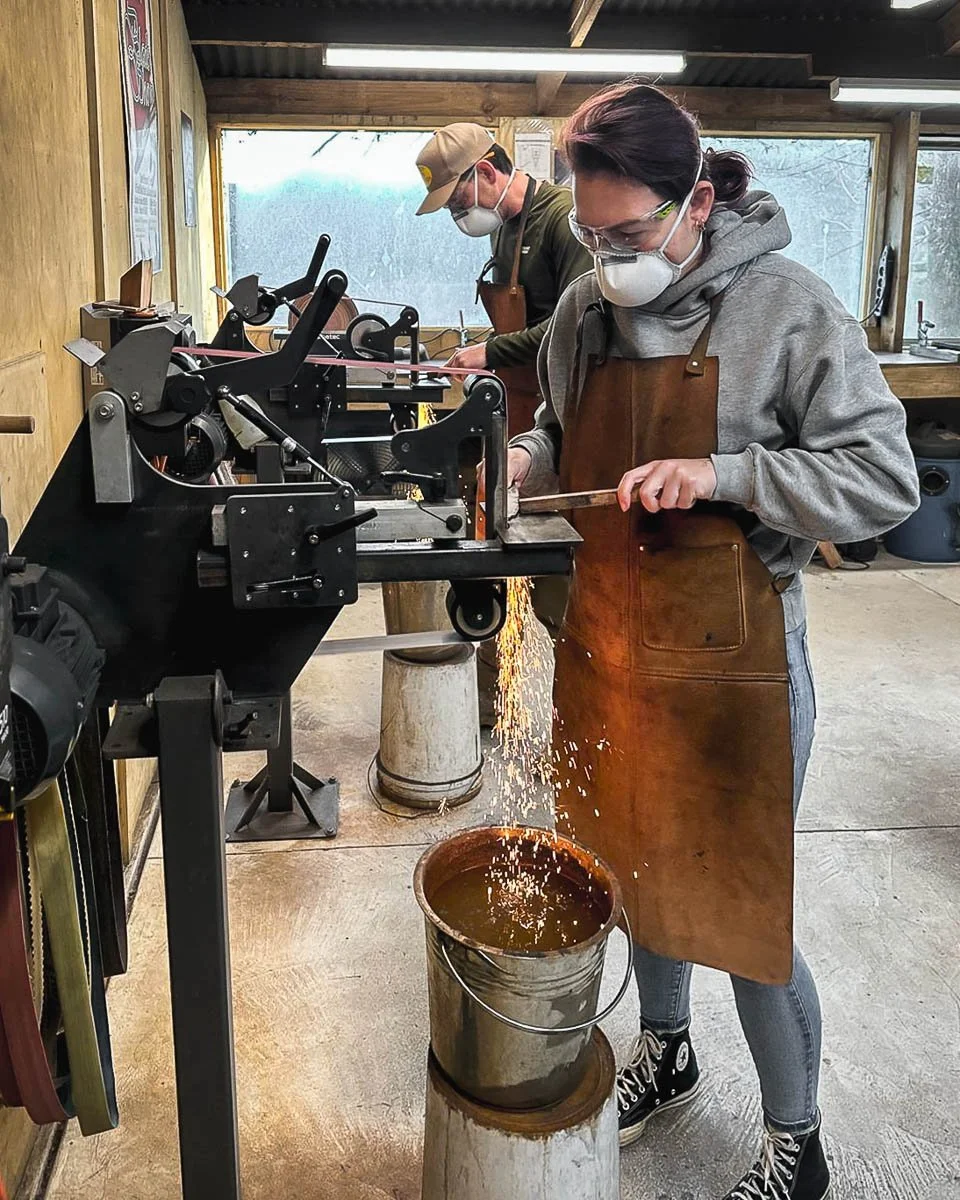 Two people wearing masks and aprons are working with a metal belt grinder in a workshop, sparks flying as they cut or grind a knife.