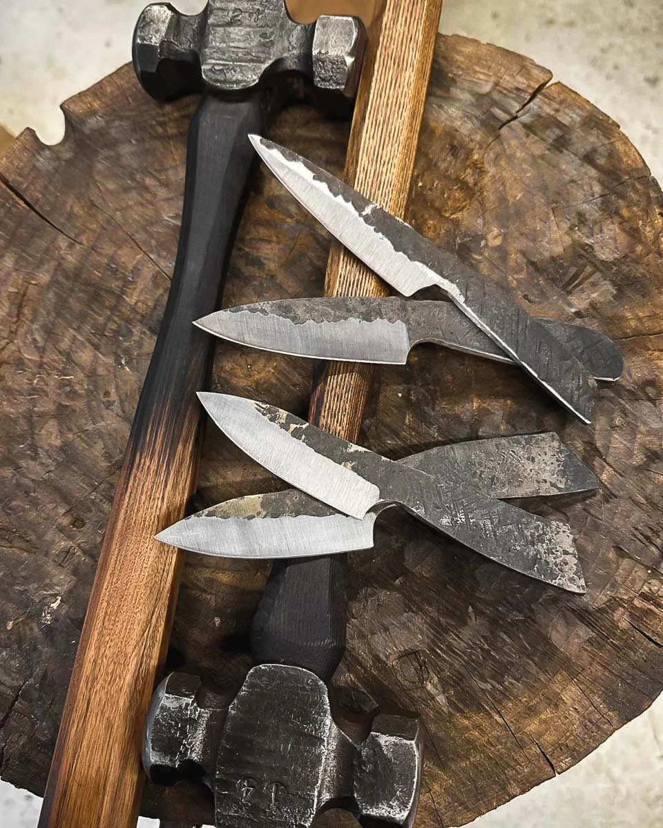 Four forged finished metal knives with rustic patterns on the blades are laid out on a wooden surface with a hammer and a chisel nearby.