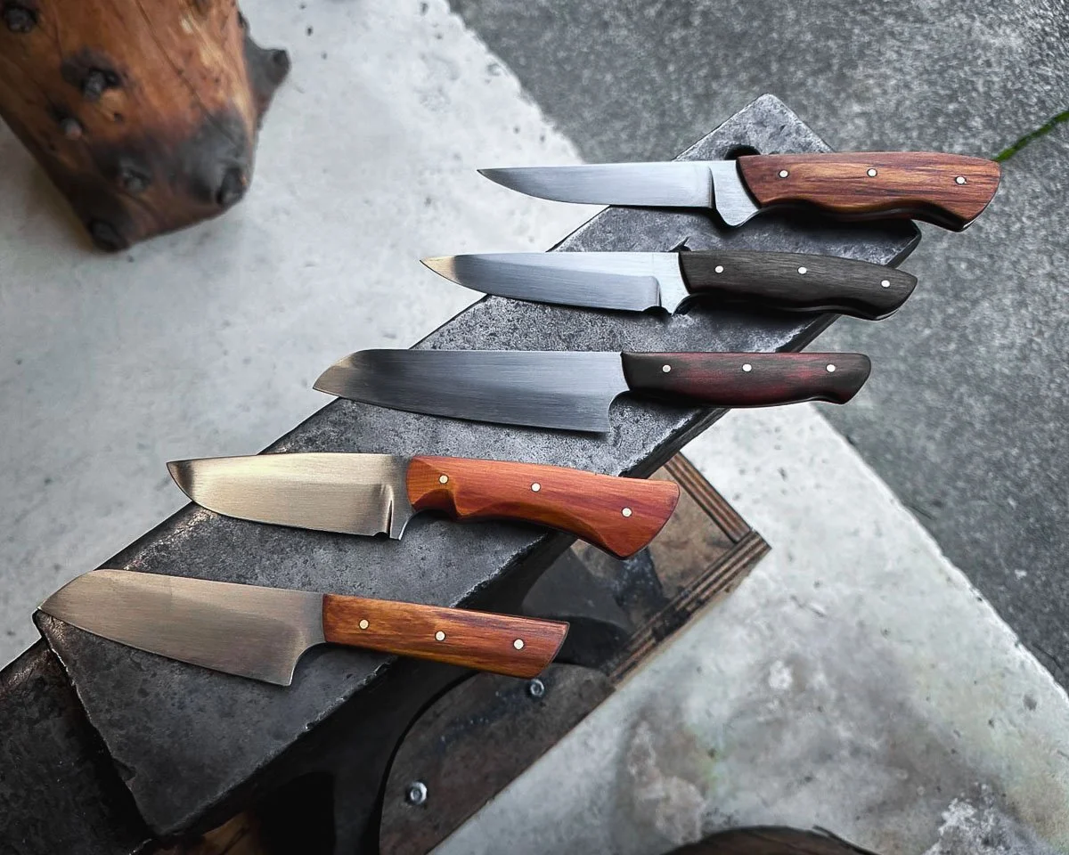 Five knives with different blade shapes and handles arranged on a rustic dark gray anvil.