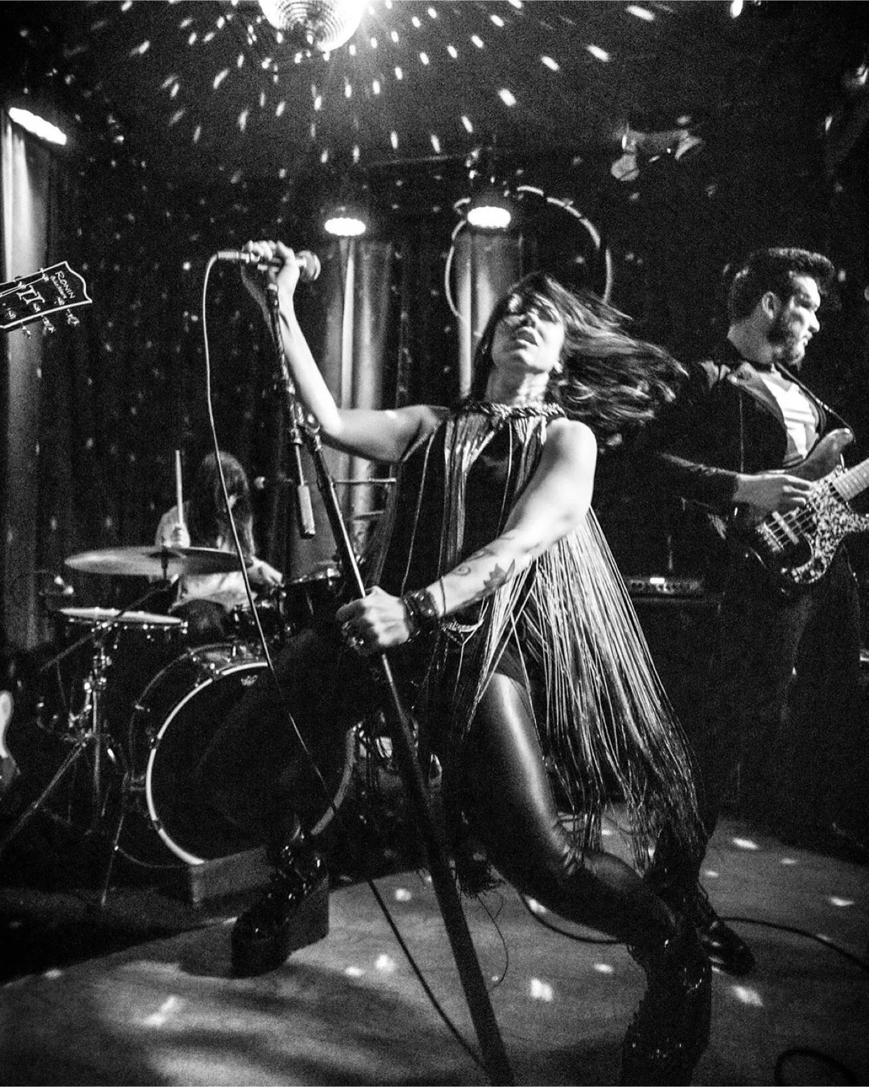 A black and white photo of a band performing on stage. The lead singer, a woman with long hair and tattoos, is holding a microphone stand and singing passionately. A drummer with long hair is playing drums in the background, and a guitarist is playing an electric guitar on the right side of the image. The stage has a star-like lighting pattern above.