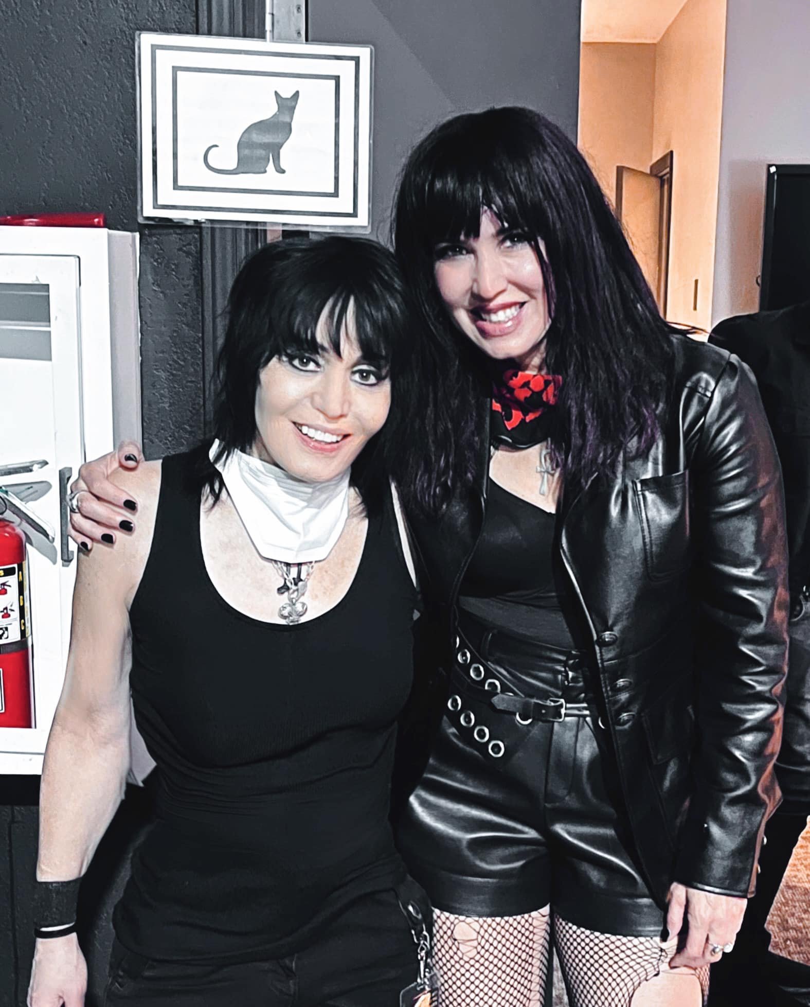 Close-up of Joan Jett and ZouZou from Soraia. One with long black hair and a leather jacket, the other with short black hair in a black dress and white scarf, both smiling and with arms around each other on a dark background with a framed cat picture