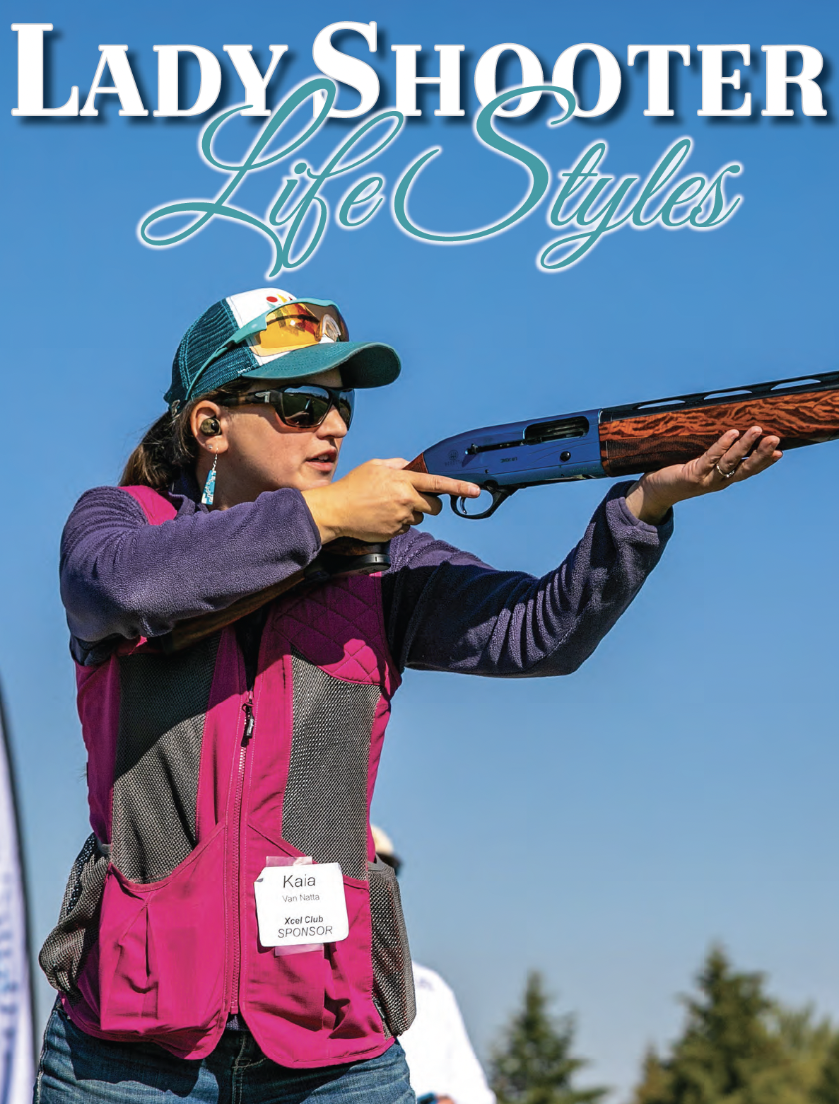 A woman wearing sunglasses, a baseball cap, and ear protection is aiming a shotgun outdoors against a clear blue sky. She is dressed in a purple jacket and a pink vest with a name tag that says 'Kaia Van Natta, Xcel Club SPONSOR'.