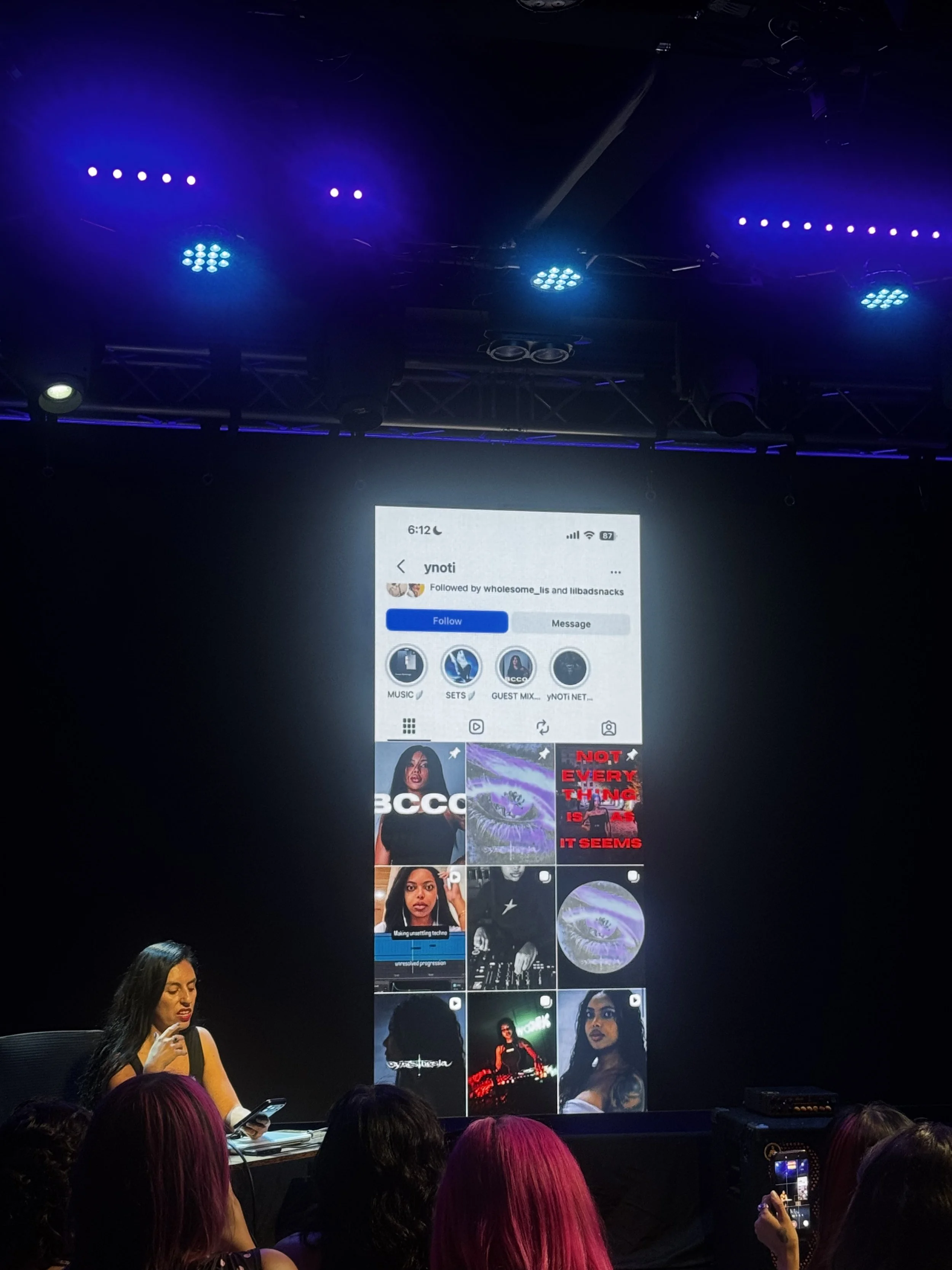 Presenter on stage holding a phone while a social media profile is projected on a large screen at a live event