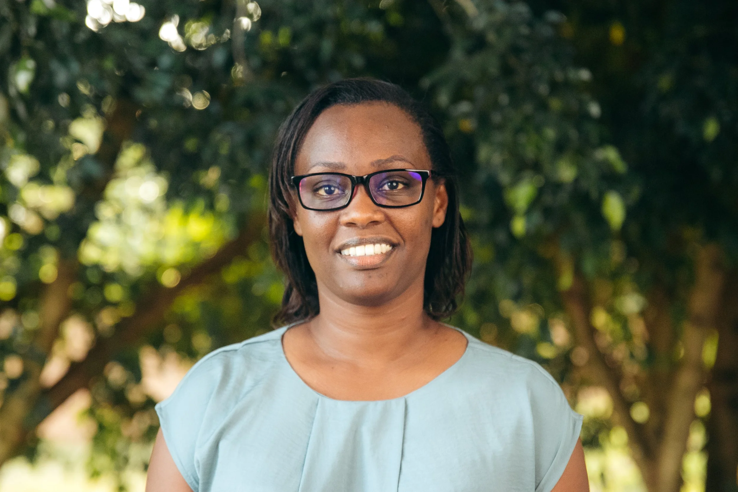 A woman with short dark hair and glasses smiling outdoors with trees in the background.