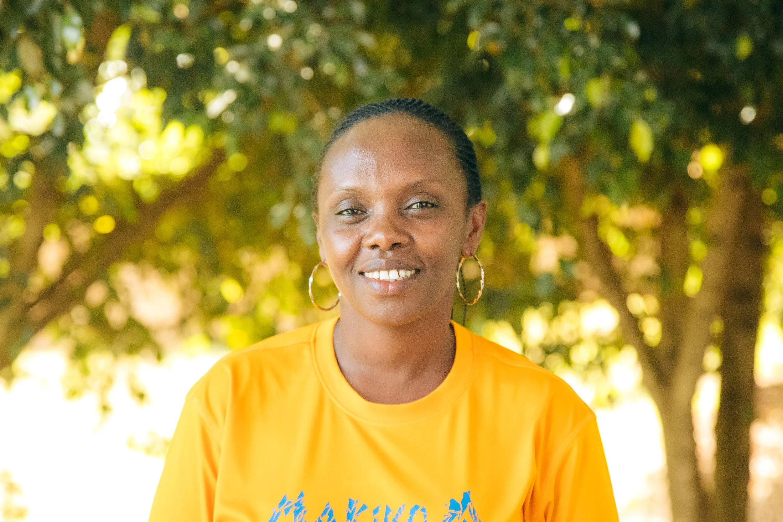 A smiling woman wearing a yellow T-shirt and hoop earrings outdoors with trees in the background.
