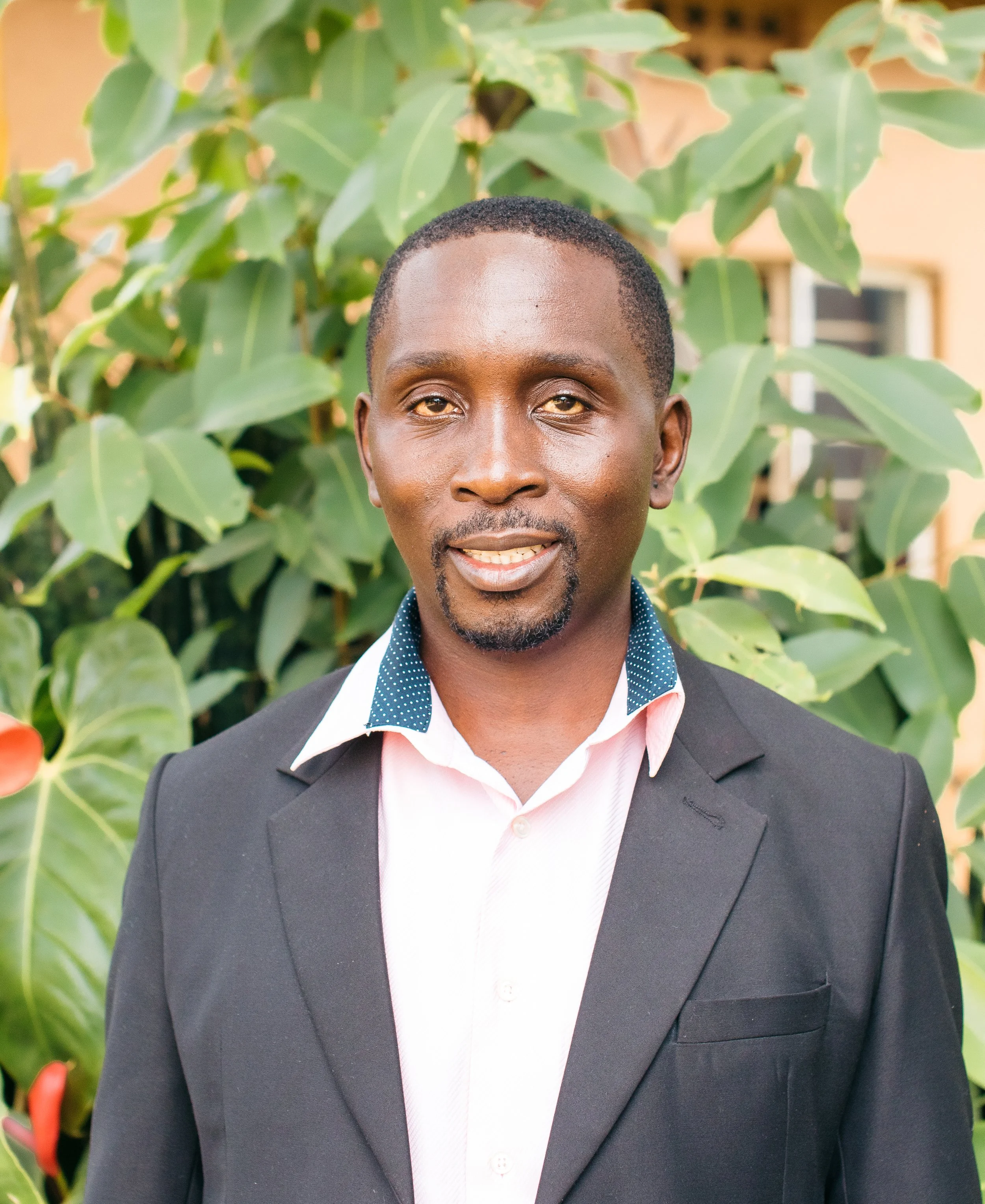 A man in a black suit jacket and white shirt with a patterned collar standing outdoors in front of green leafy plants.