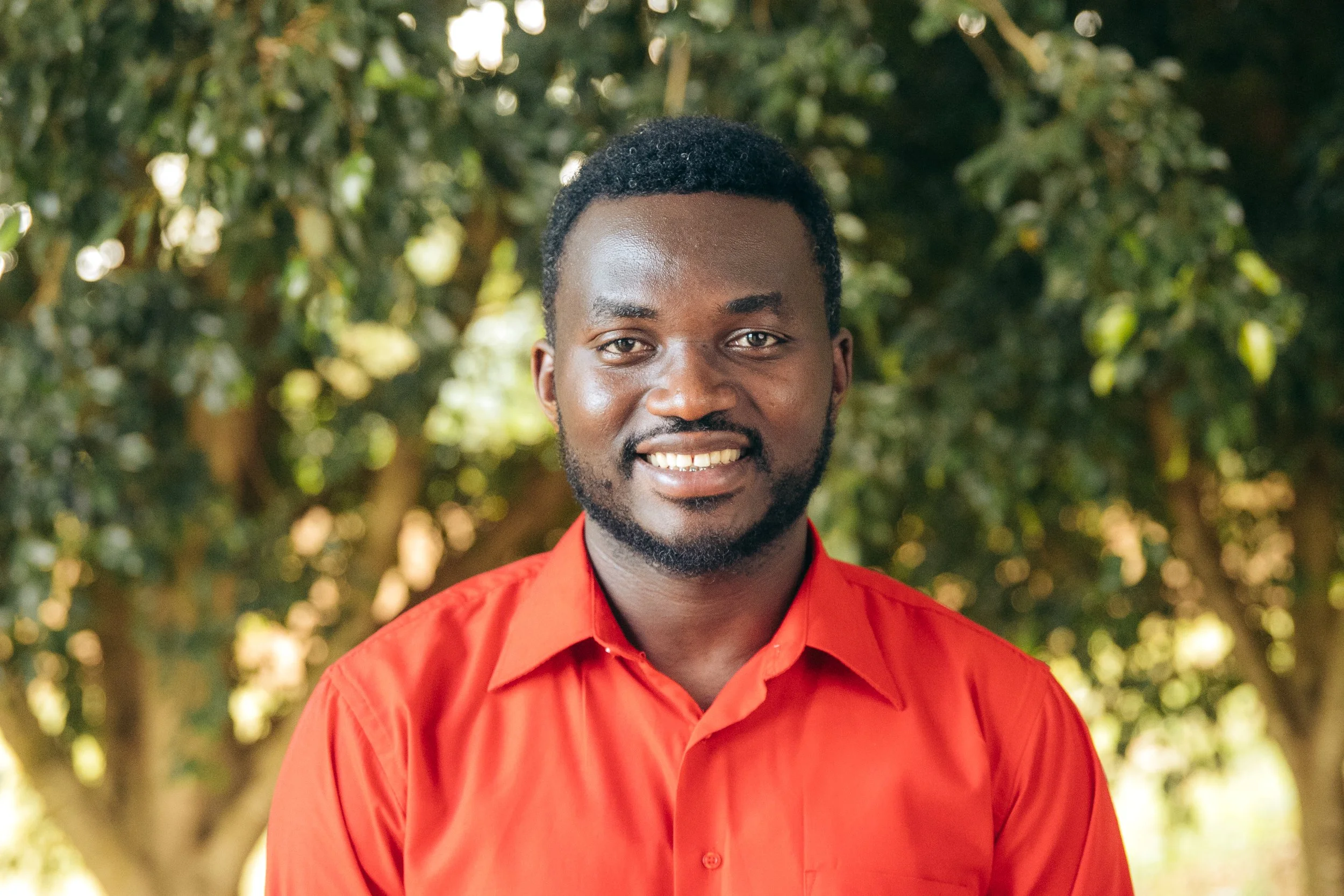 Portrait of a smiling Black man with short curly hair and a beard, wearing a red button-up shirt, outdoors with green foliage in the background.