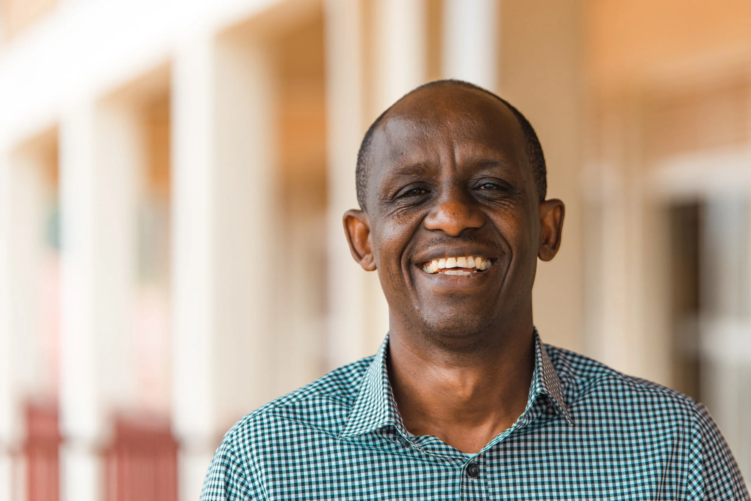 A smiling man in a checkered shirt standing in front of a blurred background of buildings or a corridor.