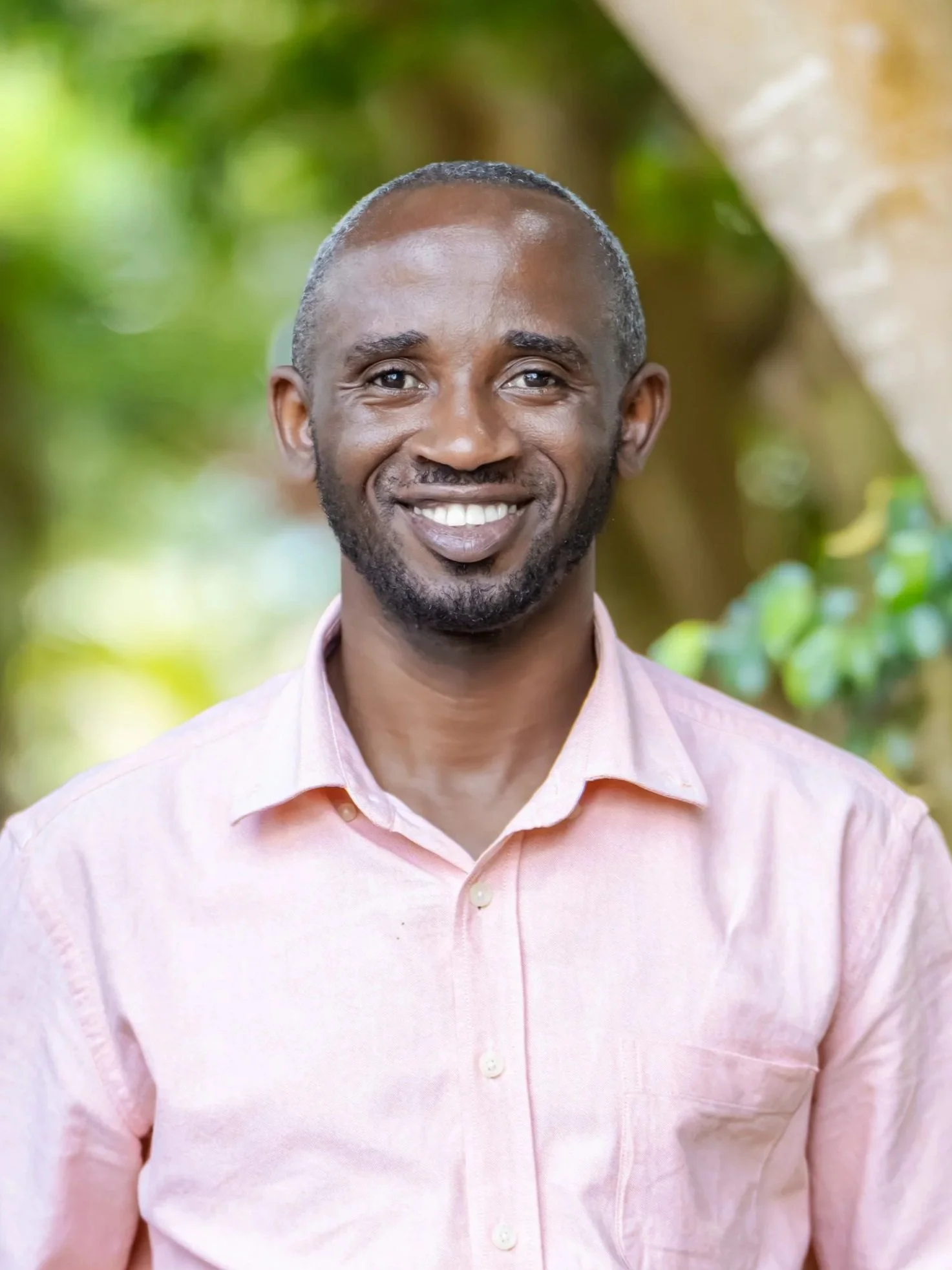 Man with short gray hair, in light pink shirt, smiling, with blurred trees in the background