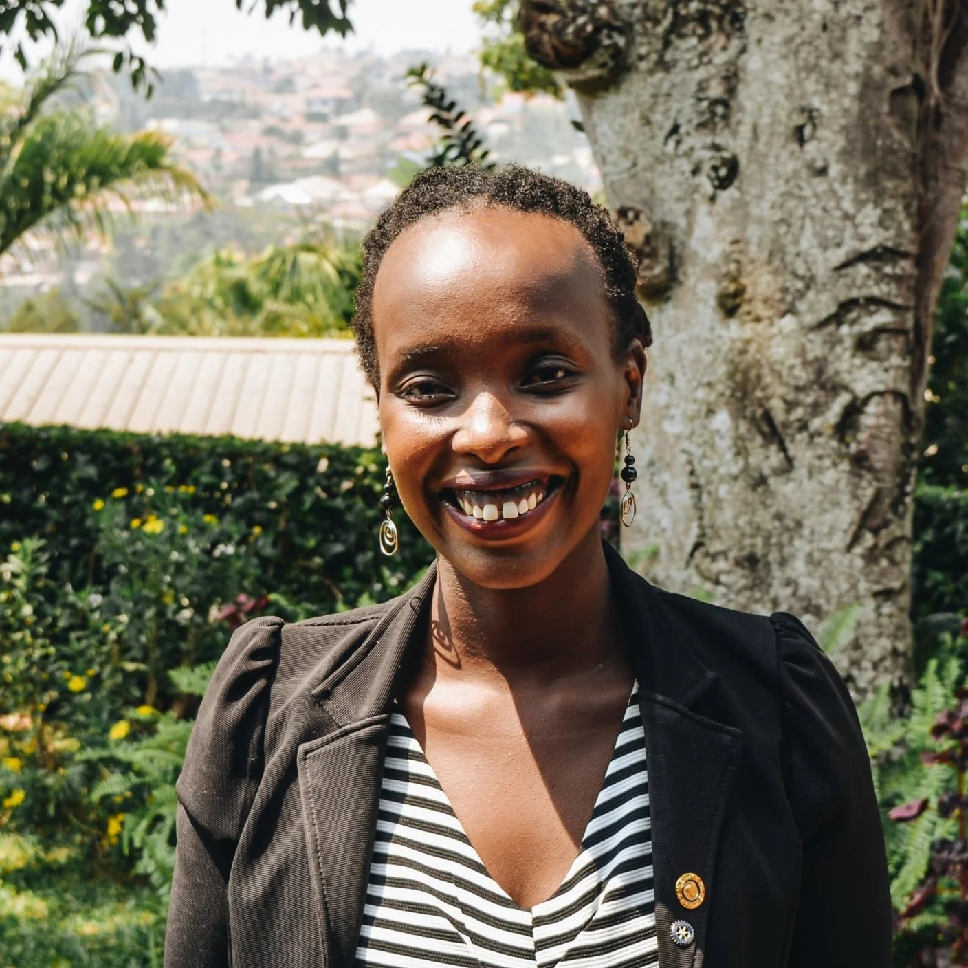 Smiling woman with short natural hair wearing a striped top and black jacket, standing outdoors in front of greenery and a large tree.