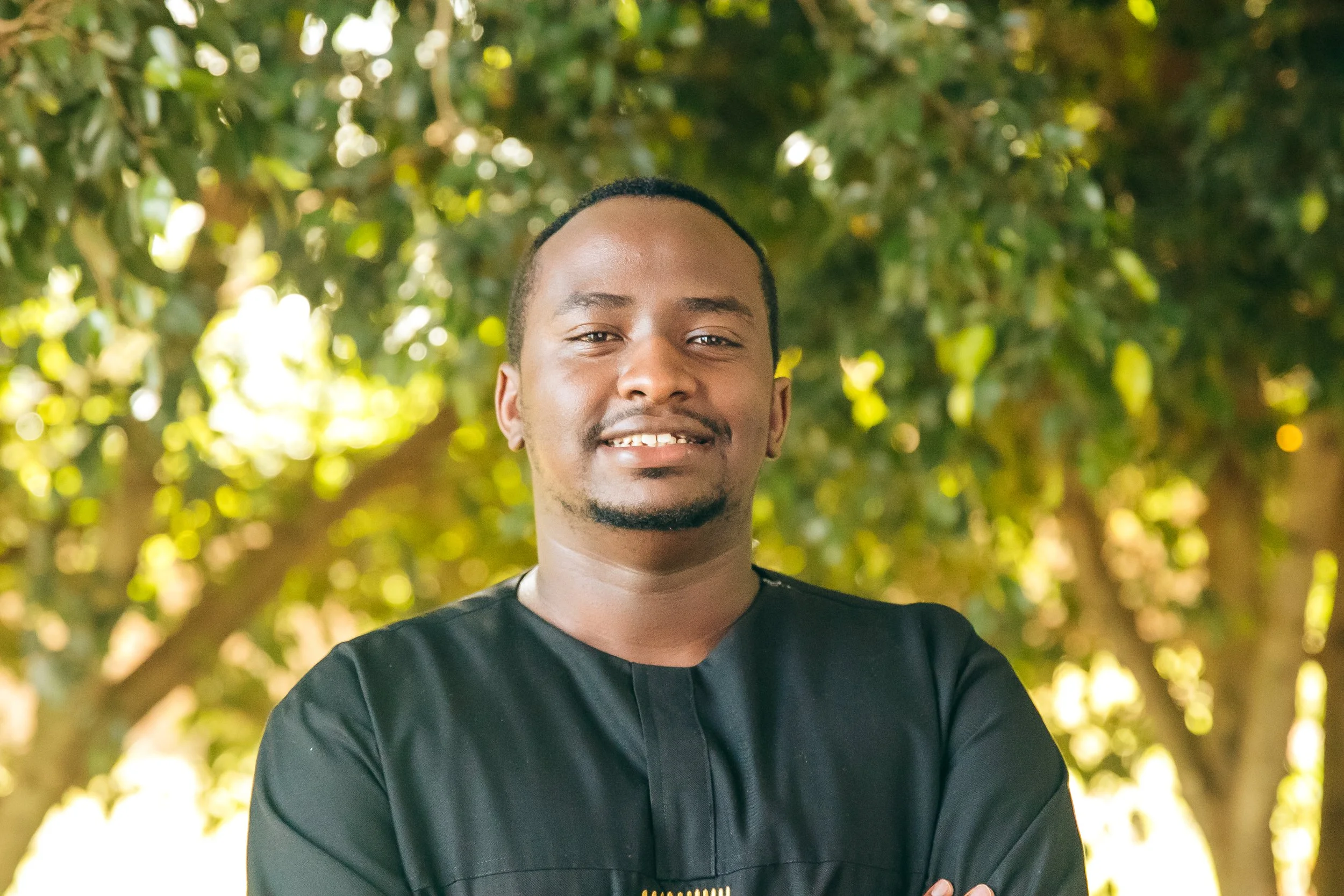 A man with dark skin, short black hair, and a mustache, wearing a black academic gown, standing outdoors with trees and bright sunlight in the background.