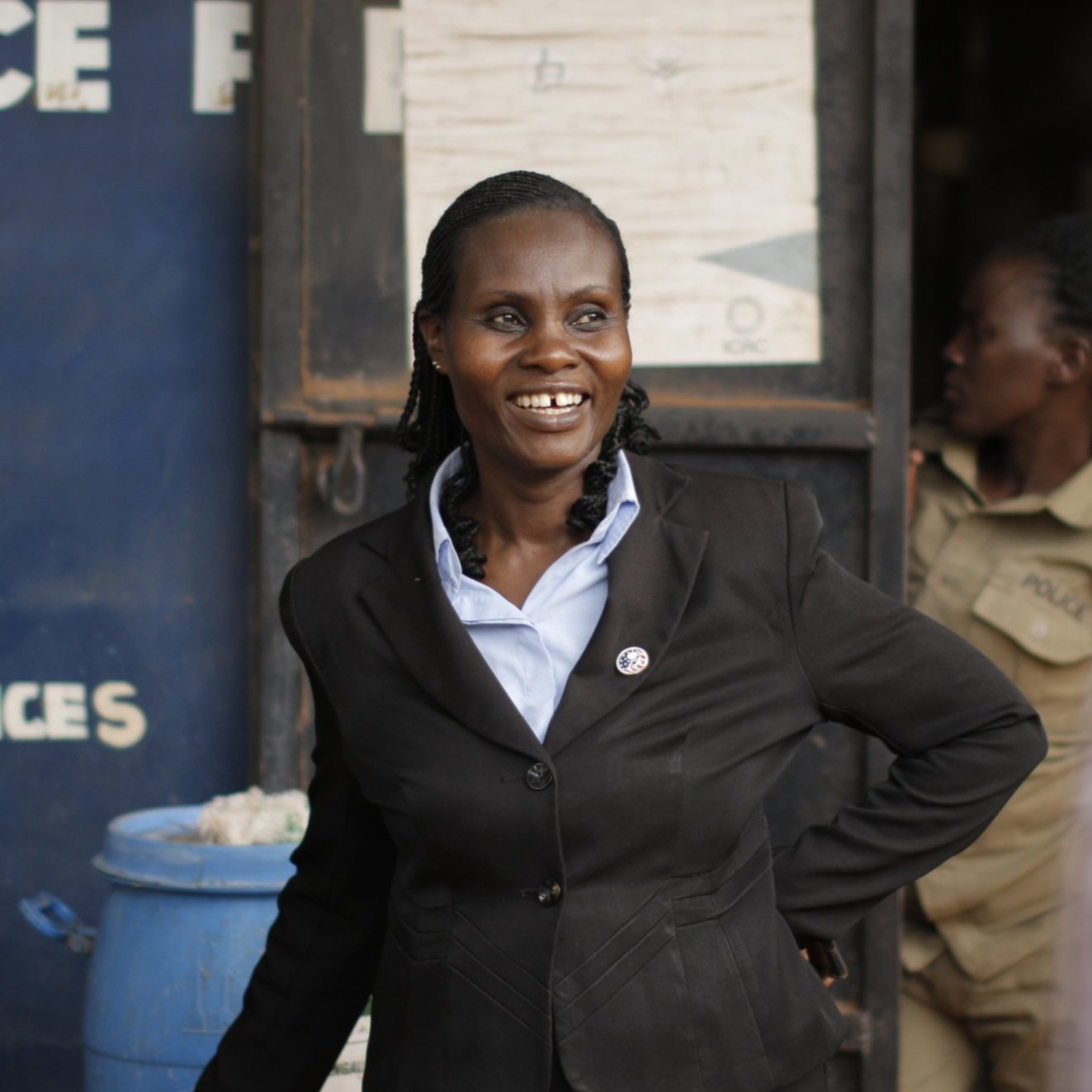 A woman in a black business suit smiling, standing in front of a blue wall and a light-colored door with a woman in a tan uniform in the background.