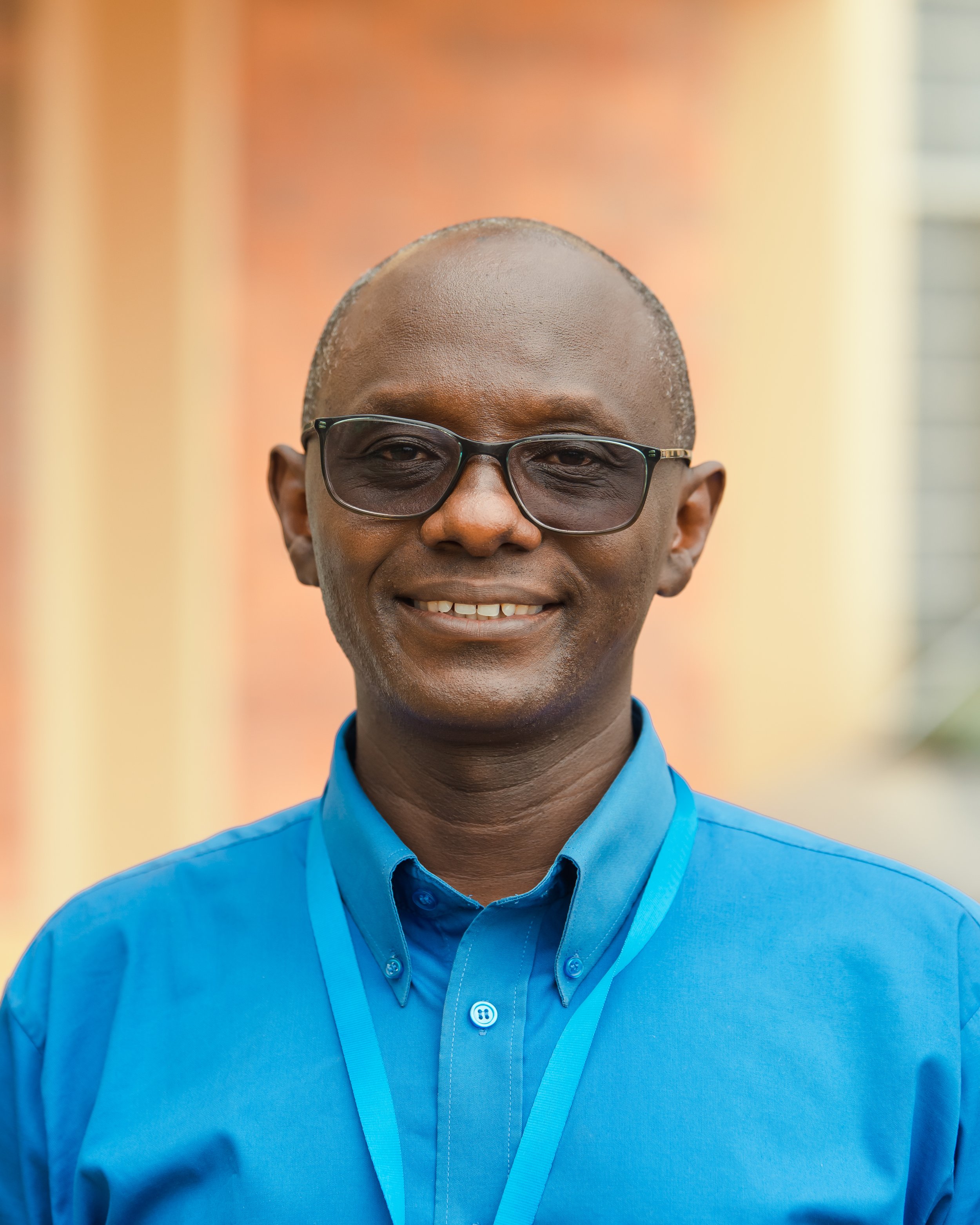 A smiling man with dark skin, wearing glasses and a bright blue collared shirt, standing outdoors with colorful blurred background.