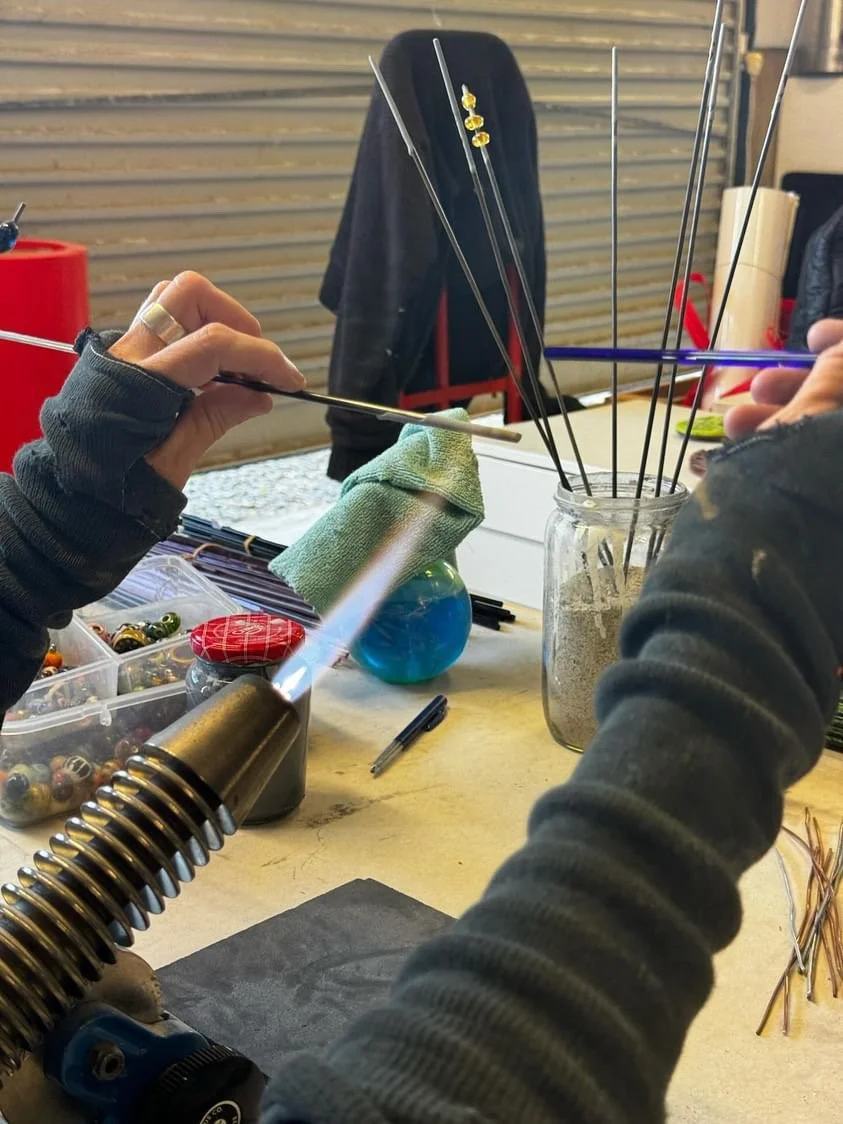 Jo Tricker on her torch, lampworking at her workbench, with tools, glass rods, and jars of glass powder on table.