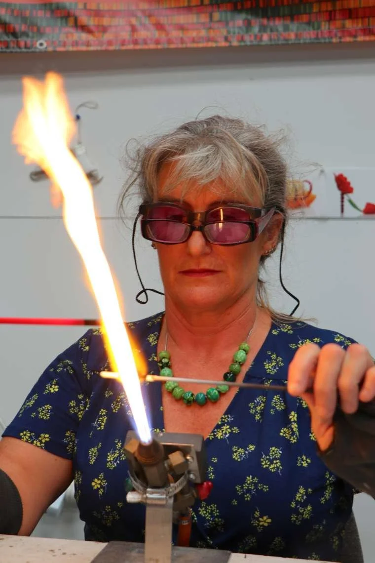 Jo Tricker,  wearing protective glasses, holding a glass rod near an open flame for lampworking.