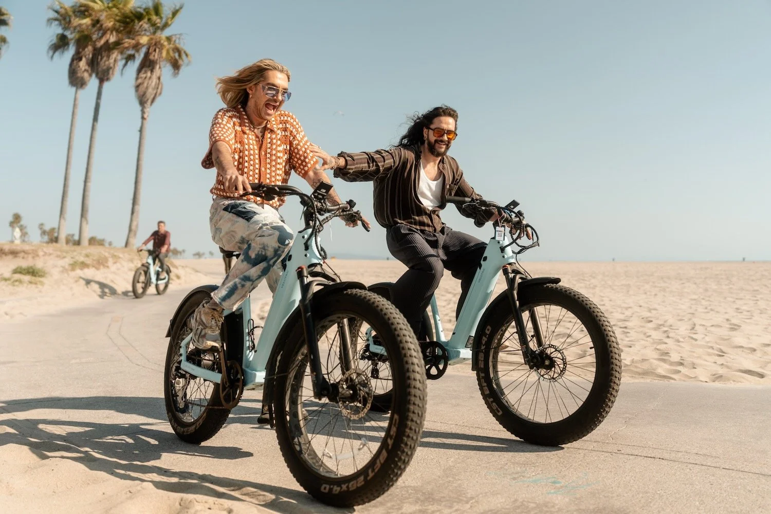 Bill and Tom Kaulitz laugh as they ride light-blue e-bikes along the beach path in Venice, Los Angeles, with palm trees behind them.