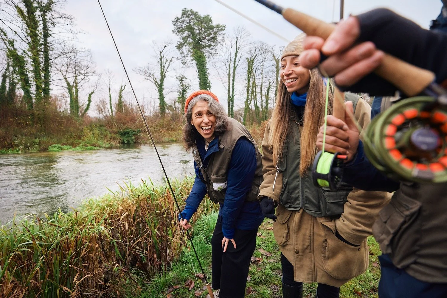 Two women in woolly hats and fishing vests laughing on the grassy bank of the River Test during a fly-fishing lesson.