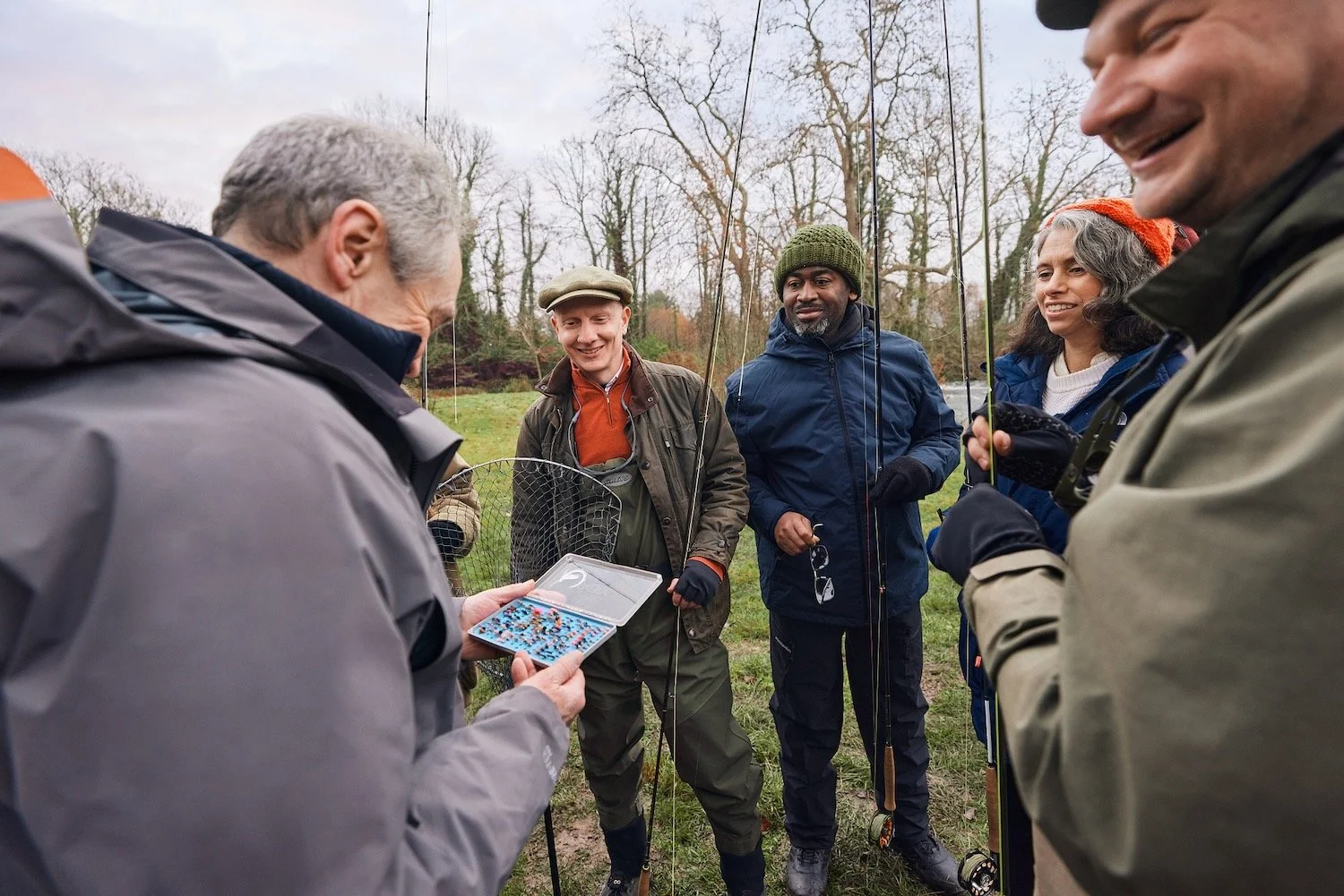Paul Whitehouse showing an open box of fishing flies to a smiling group of guests holding fly rods and a landing net on the bank of the River Test.