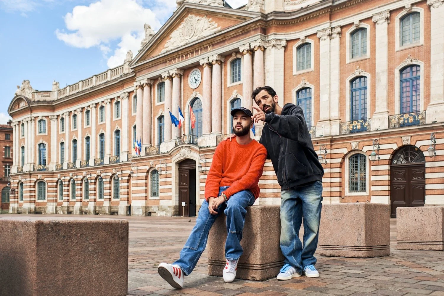 BigFlo & Oli lean on a stone block on Place du Capitole in Toulouse, with the Capitole's pink brick facade behind them.