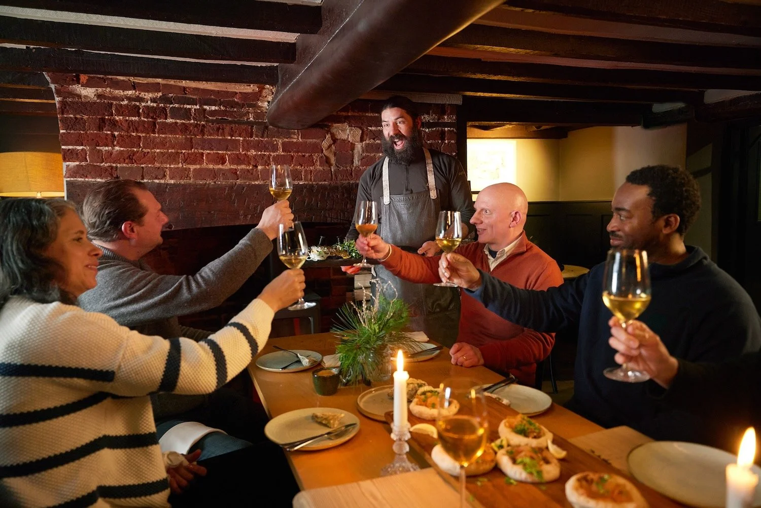 Chef Nick Weston serving fire-cooked trout to Paul Whitehouse and guests at a candlelit table inside The Greyhound on the Test pub in Stockbridge.