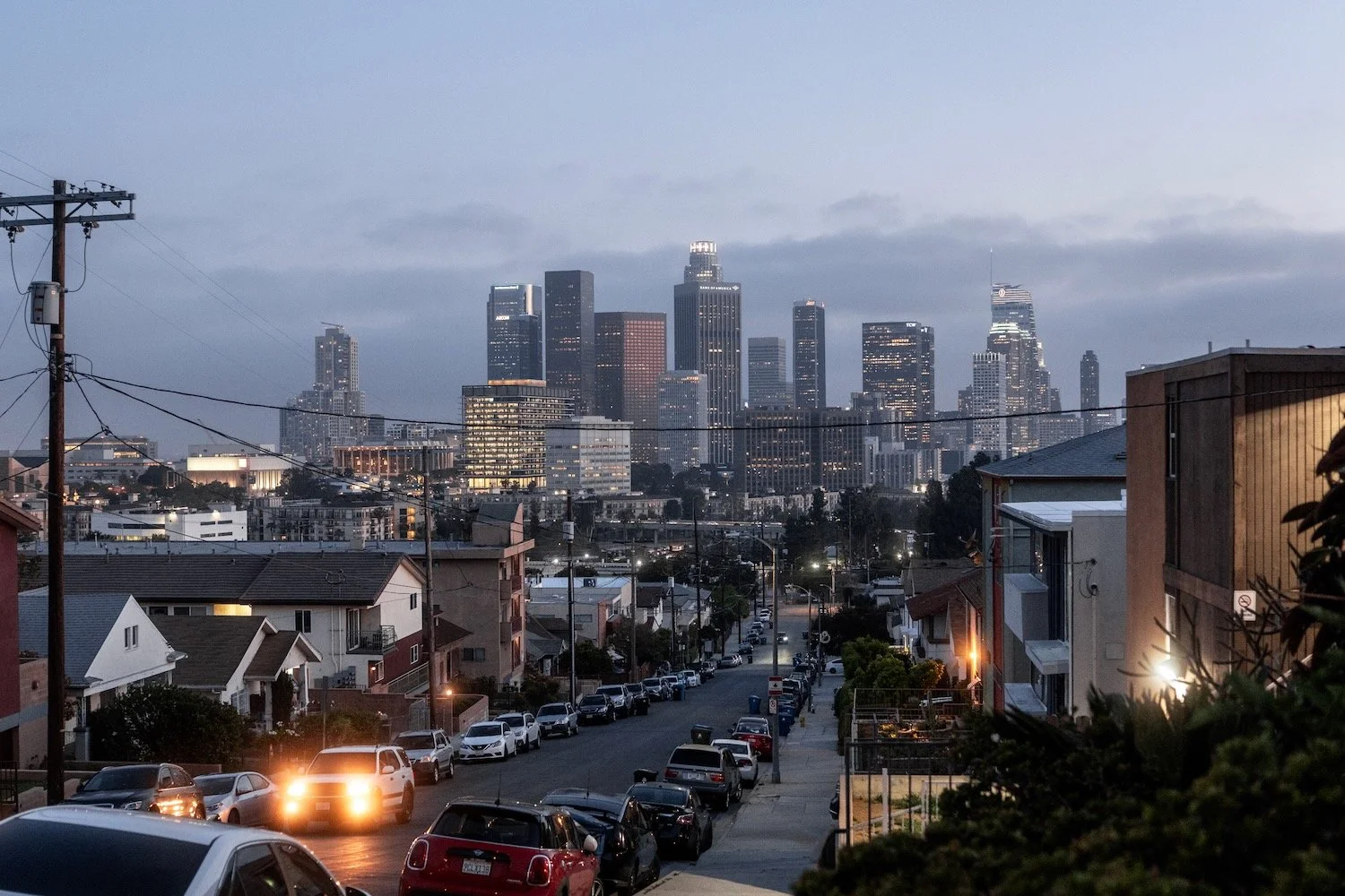Downtown Los Angeles skyline at dusk, seen from a hilltop street in Figueroa Terrace, with car headlights on the road below.