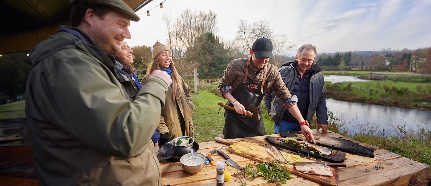 Chef Nick Weston nailing a trout fillet to a wooden plank with a hatchet while other guests watch, at an outdoor table beside the River Test.