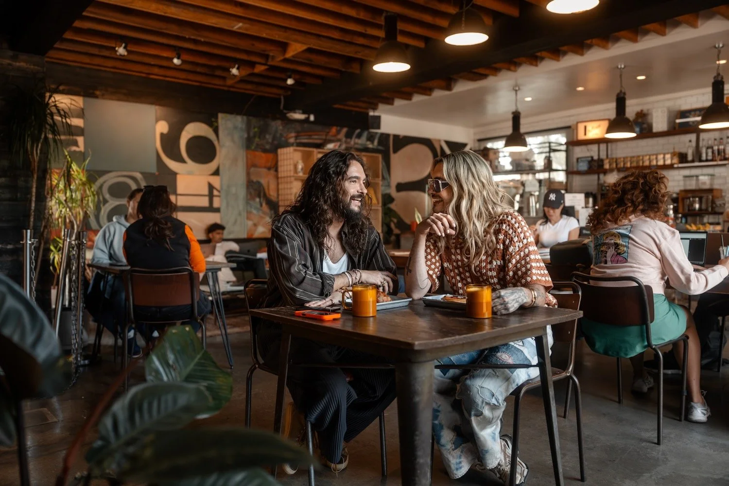 Bill and Tom Kaulitz laugh over coffee at a table in Deus Ex Machina, a busy café in Venice, Los Angeles.
