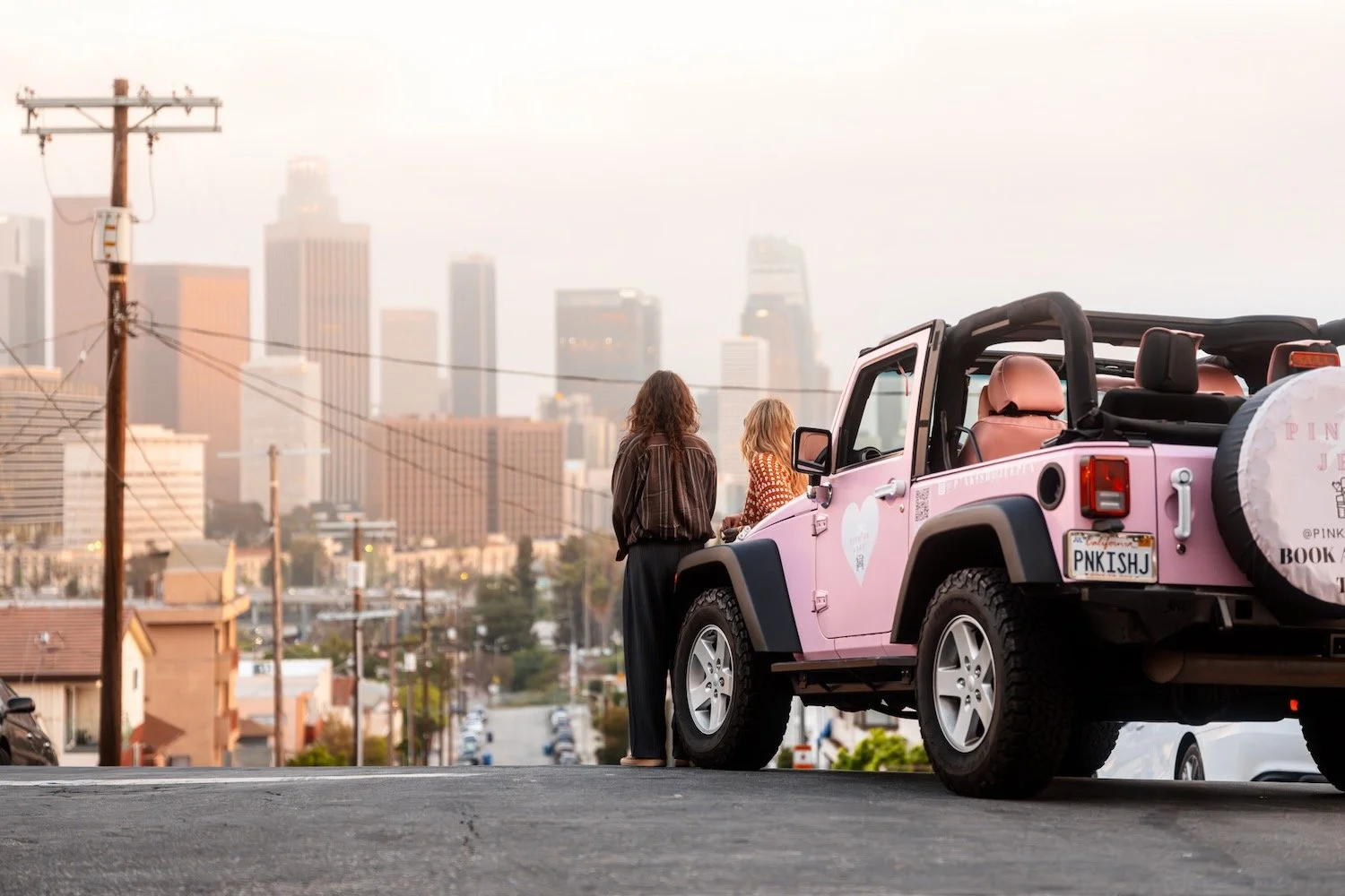 Bill and Tom Kaulitz stand beside a pink Jeep on a hilltop street, looking out over the downtown Los Angeles skyline.