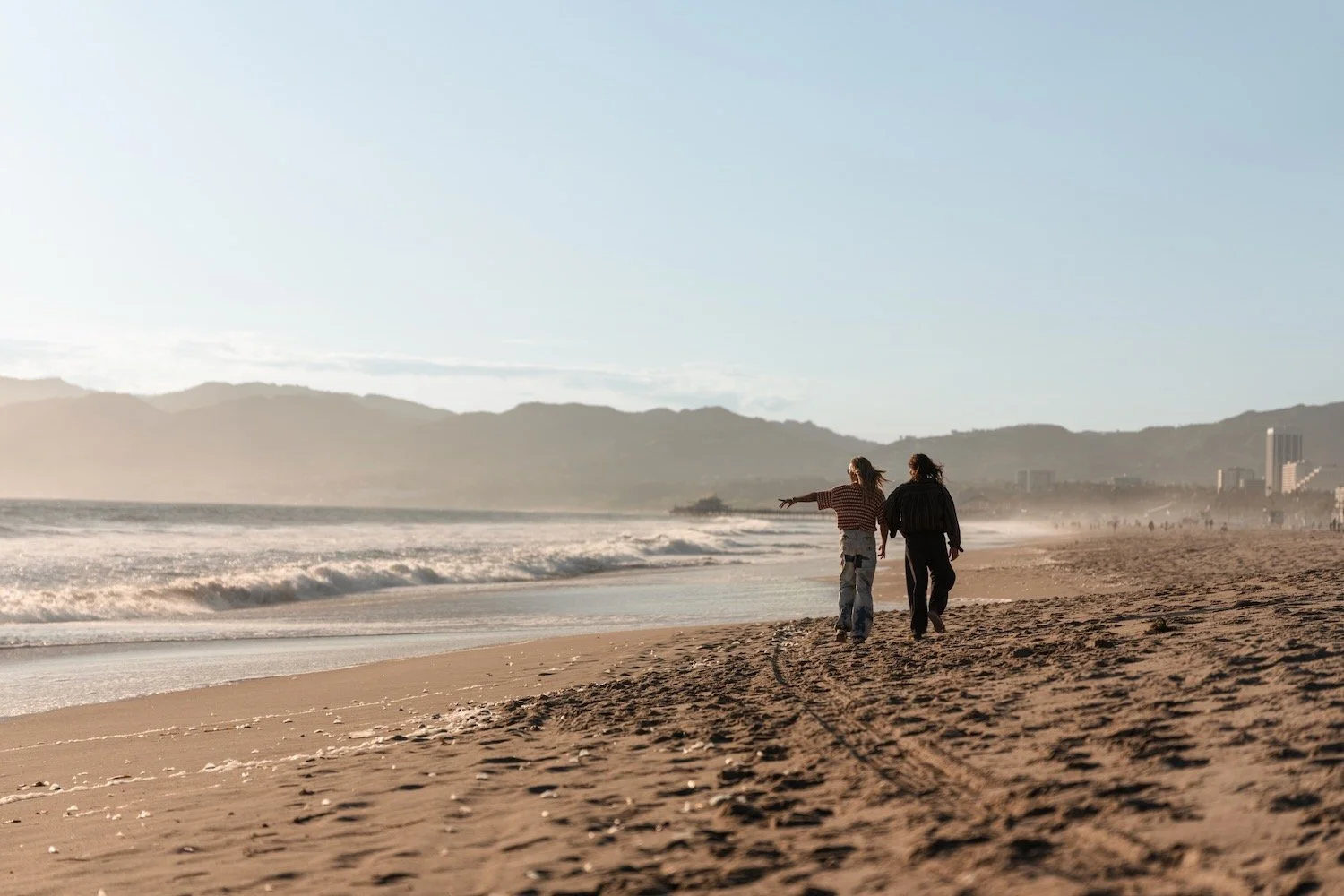 Bill and Tom Kaulitz walk along the shoreline at Venice Beach in Los Angeles, with waves and mountains in the distance.