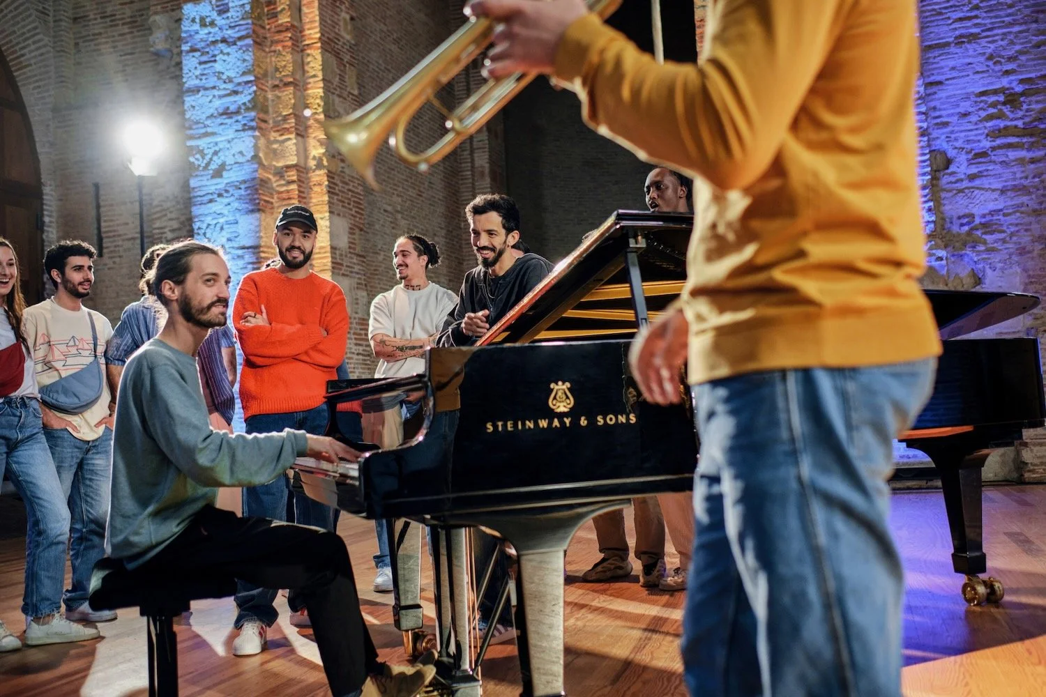 BigFlo & Oli and travelers gather around a grand piano on the stage of the Auditorium Saint-Pierre-des-Cuisines.