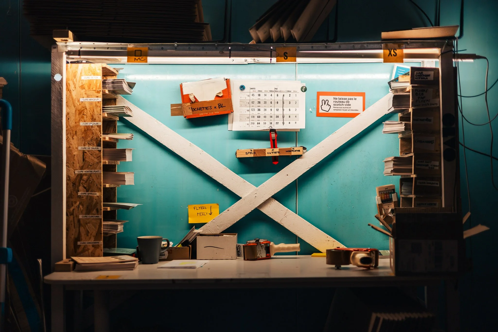 A workbench with shelves holding papers, tools, and office supplies against a teal wall with various signs and notes, including a calendar and colored labels.