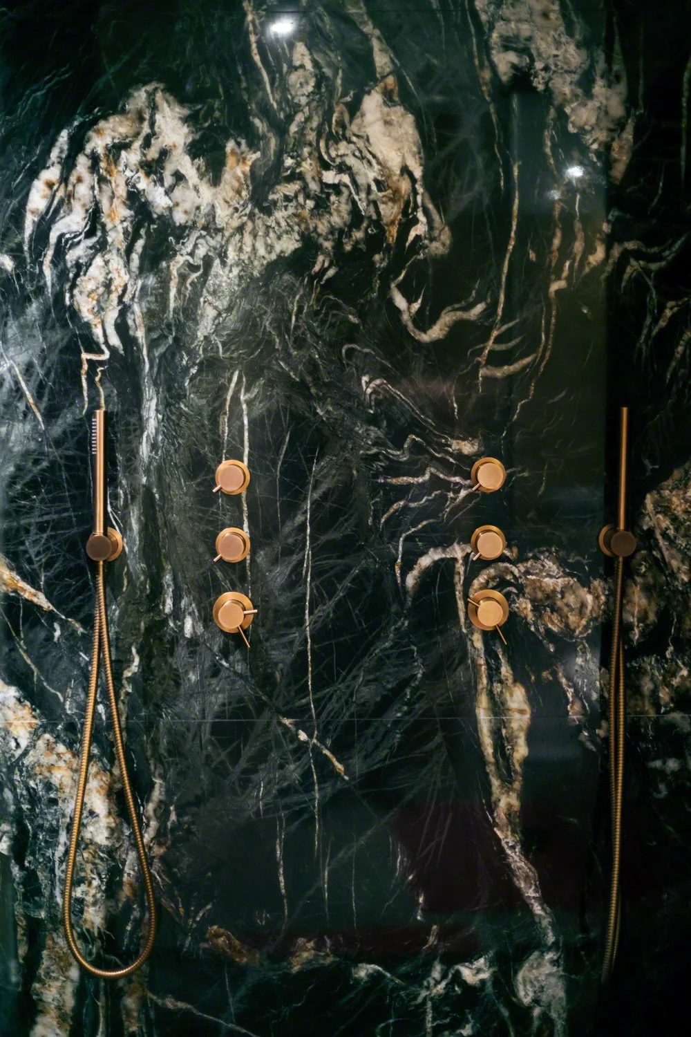 A modern shower wall made of black marble with white and gold veining, equipped with multiple gold shower controls and handheld shower heads.