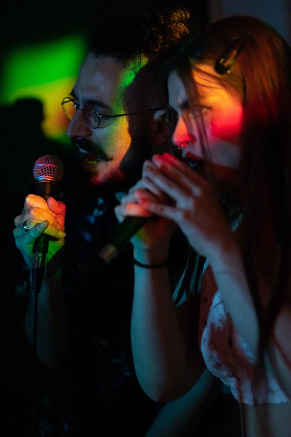 Two people singing into microphones, illuminated by colorful stage lights, in a dimly lit environment.