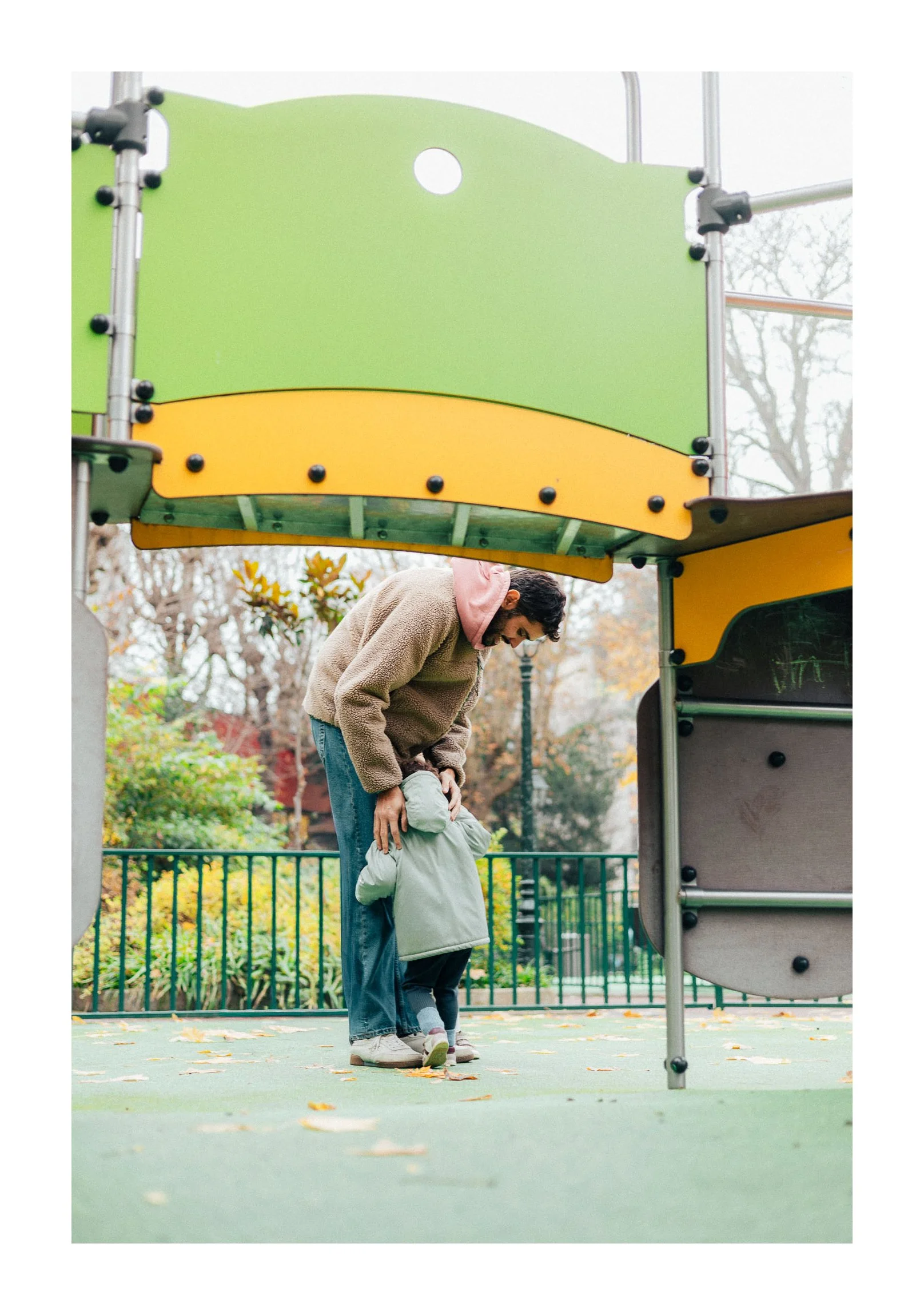 A man helping a child in a green jacket on an outdoor playground with fall leaves on the ground and trees in the background.