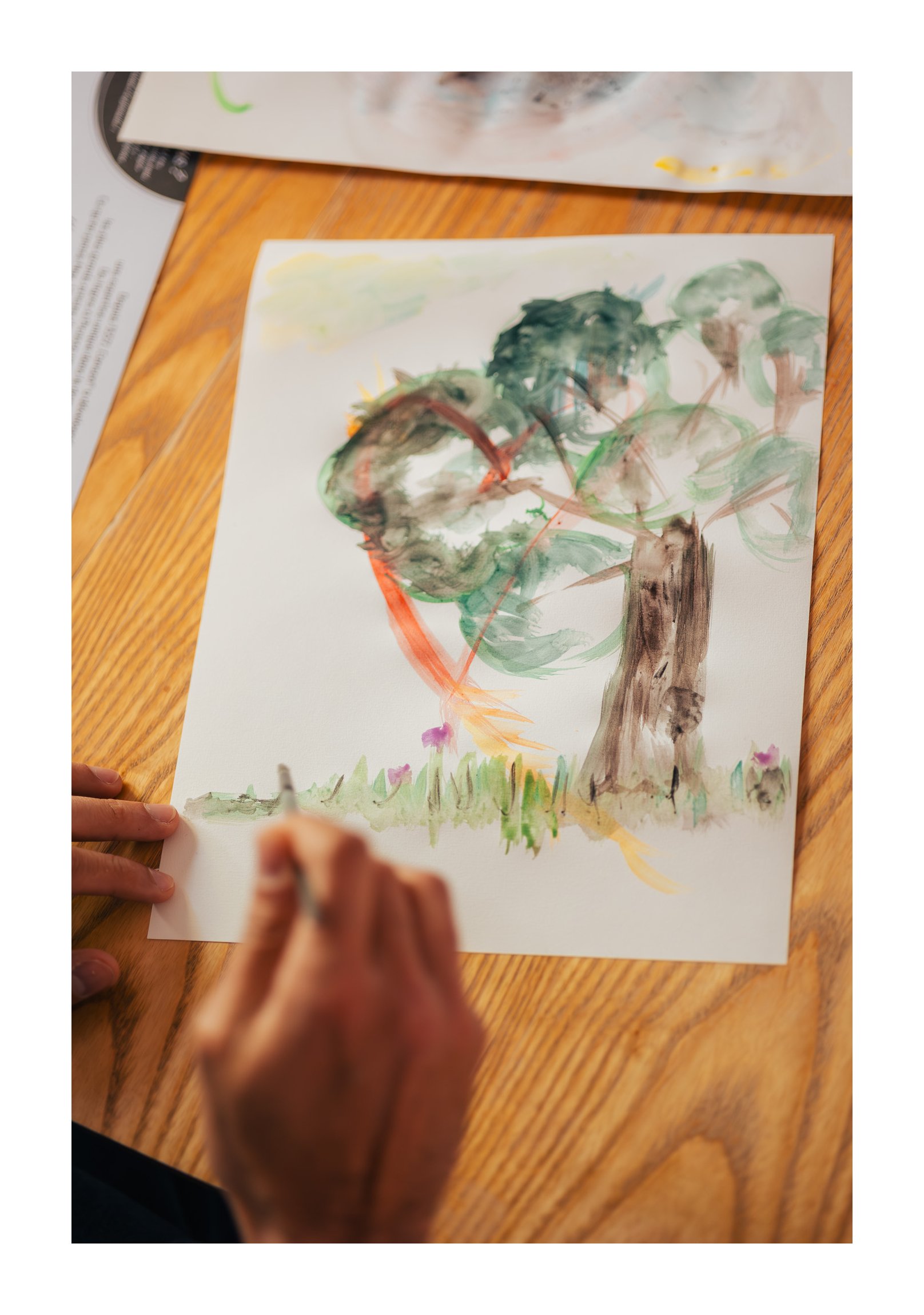 Child's hand coloring a watercolor picture of a large tree with green leaves and a brown trunk on a white sheet of paper, on a wooden table.