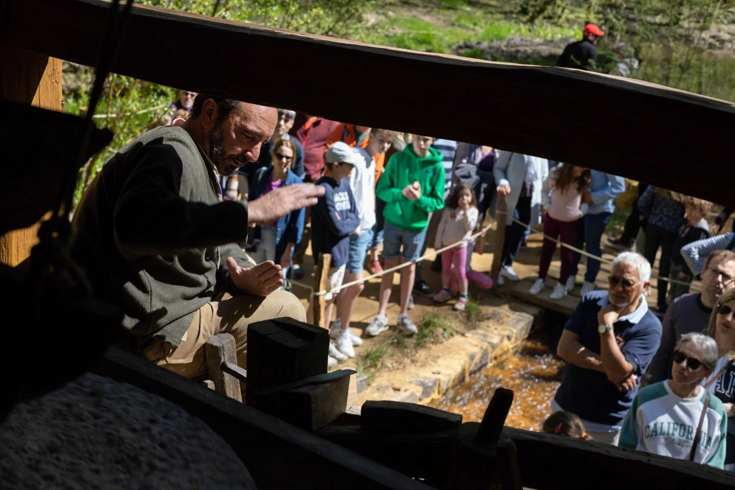 A man performing a demonstration or presentation in front of a crowd in an outdoor setting near a small stream, viewed from beneath a structure.