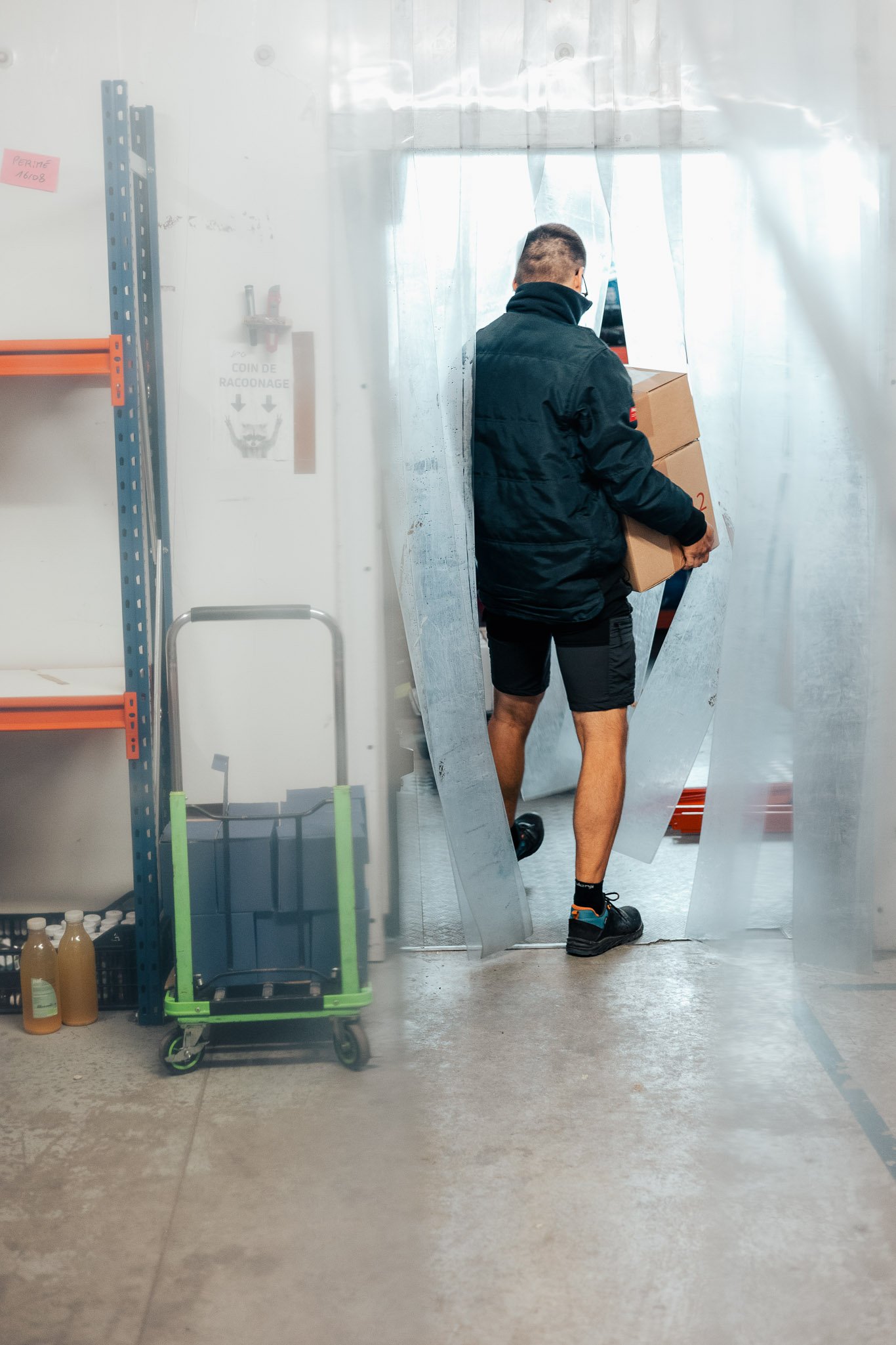 A man carrying a box through a curtain into a room, viewed from behind, with shelves and bottles on the left side.