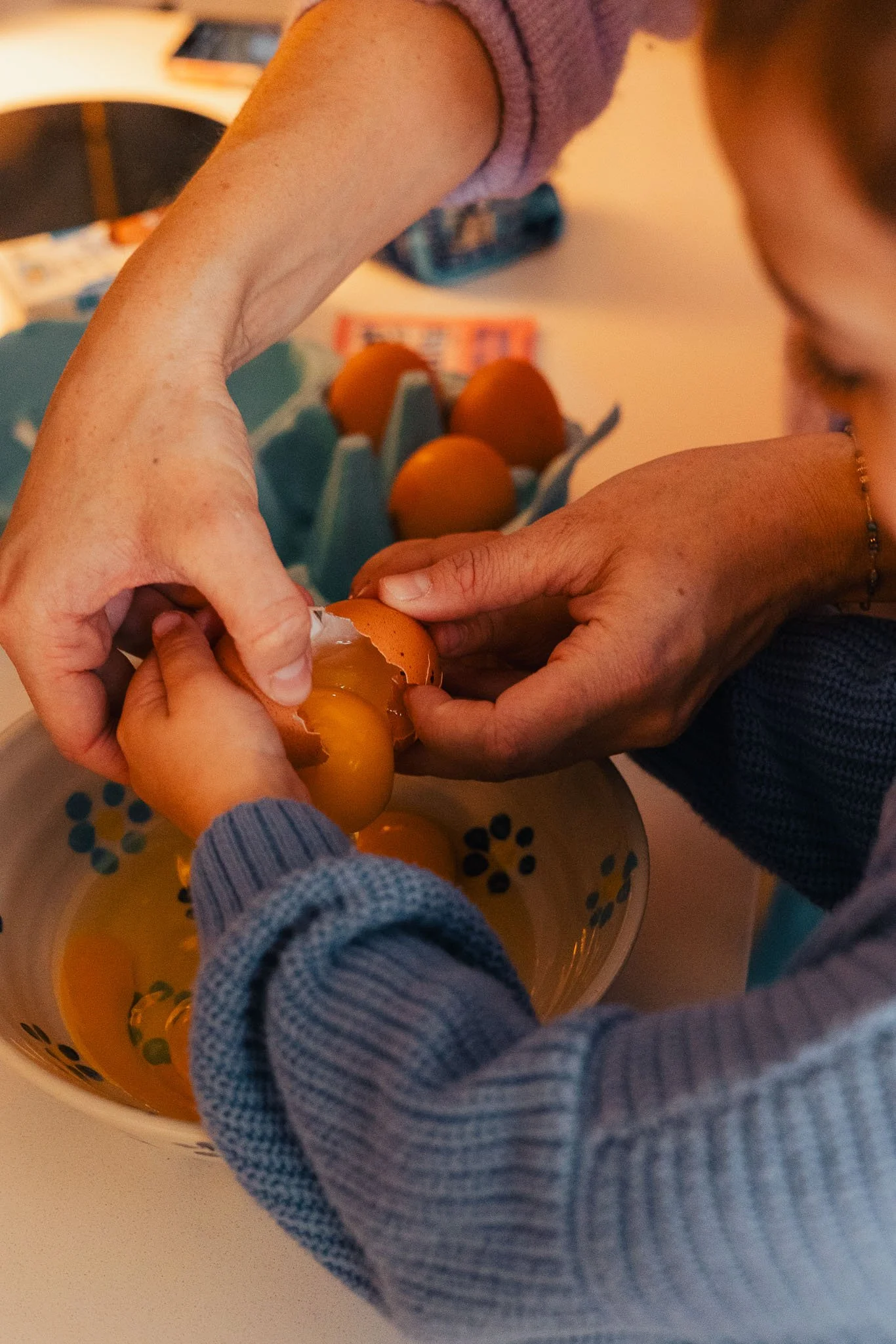 A person helping a child crack an egg into a bowl, with several eggs in an egg carton on the table.