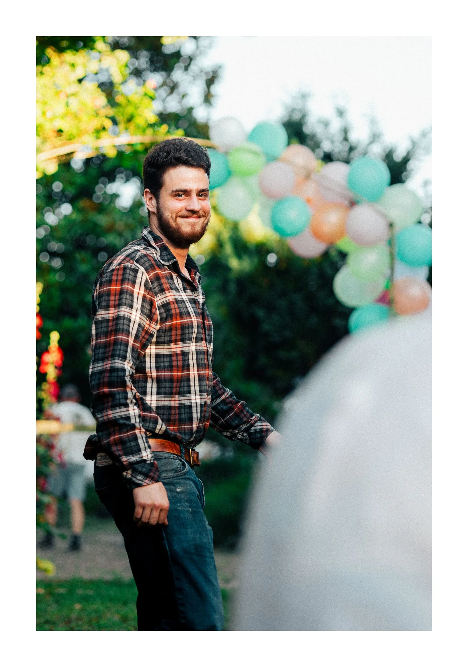 A man with dark hair and a beard, smiling, wearing a plaid shirt and jeans, standing outdoors near a colorful balloon arch at a festive event.