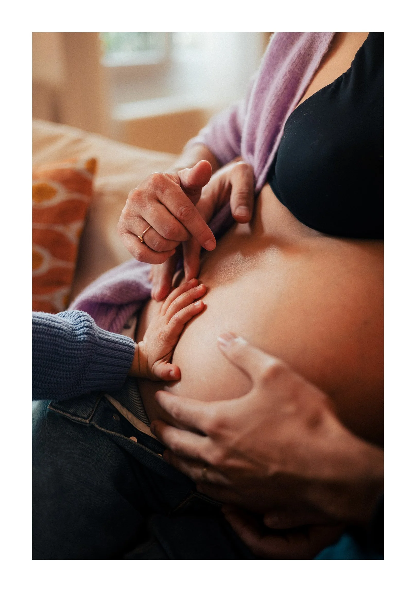 A woman is lying down with a child touching her pregnant belly, and another hand is gently holding her stomach.