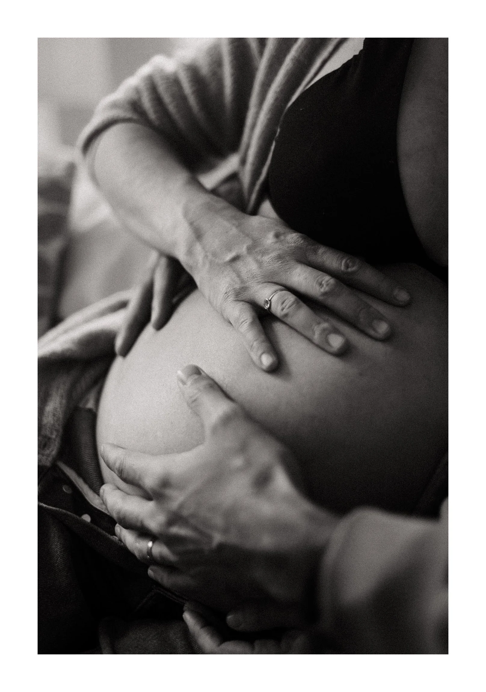 Black and white photo of a person with a ring on their finger, gently touching their pregnant belly with their hand, while another hand, possibly a partner's, is also placed on the belly, symbolizing affection and connection.