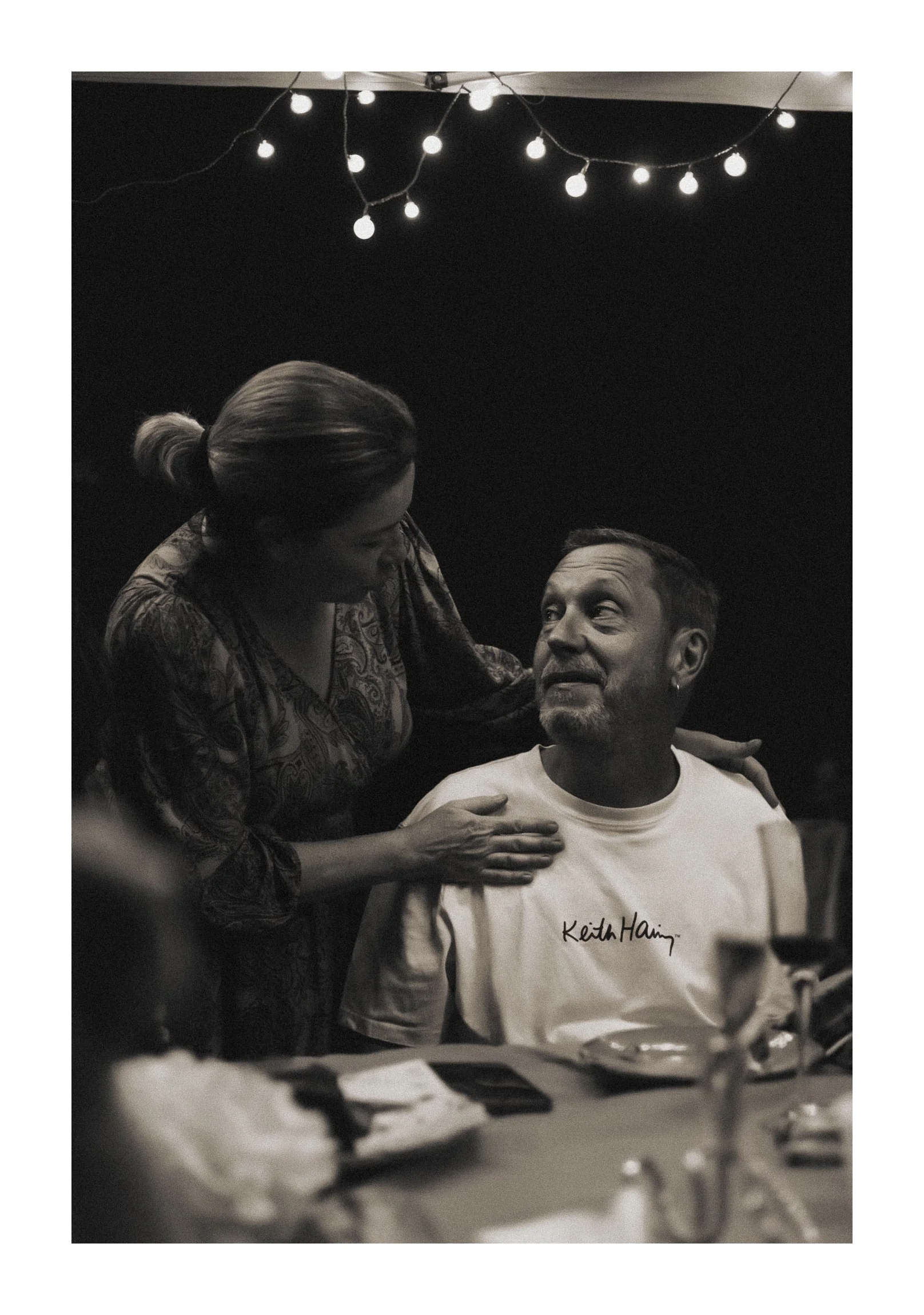 A woman is leaning over a man who is seated at a table, touching his shoulder and engaging in conversation. The man has short hair, a goatee, and is wearing a white t-shirt with the signature "Keith Haring" on it. String lights are hanging above them