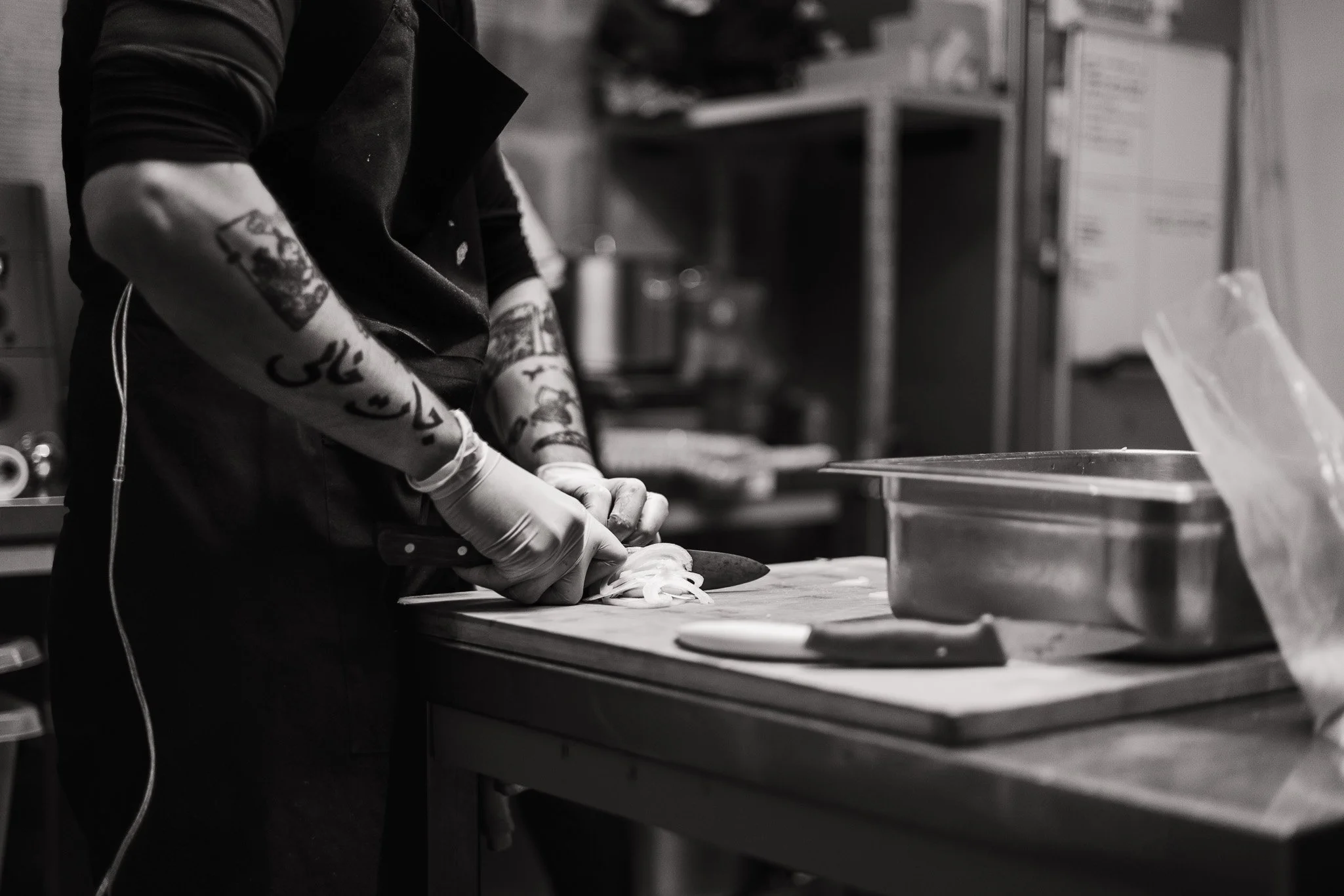 A tattooed person wearing gloves is chopping onions on a cutting board in a kitchen.