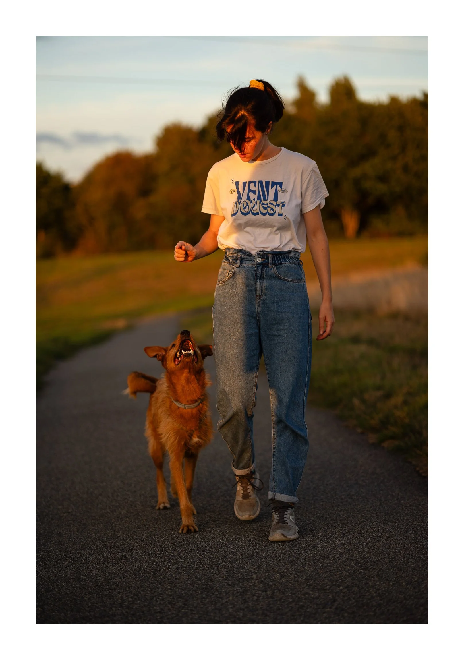 A young woman walking with a golden retriever dog on a paved path outdoors during sunset, with trees in the background.